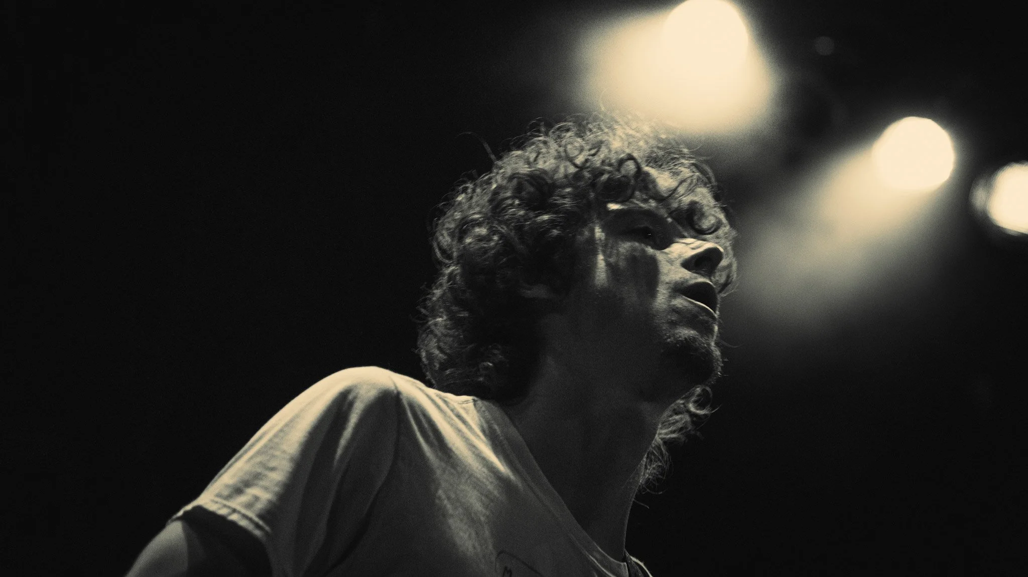 A black and white photo of a person with curly hair singing or speaking passionately under stage lights.