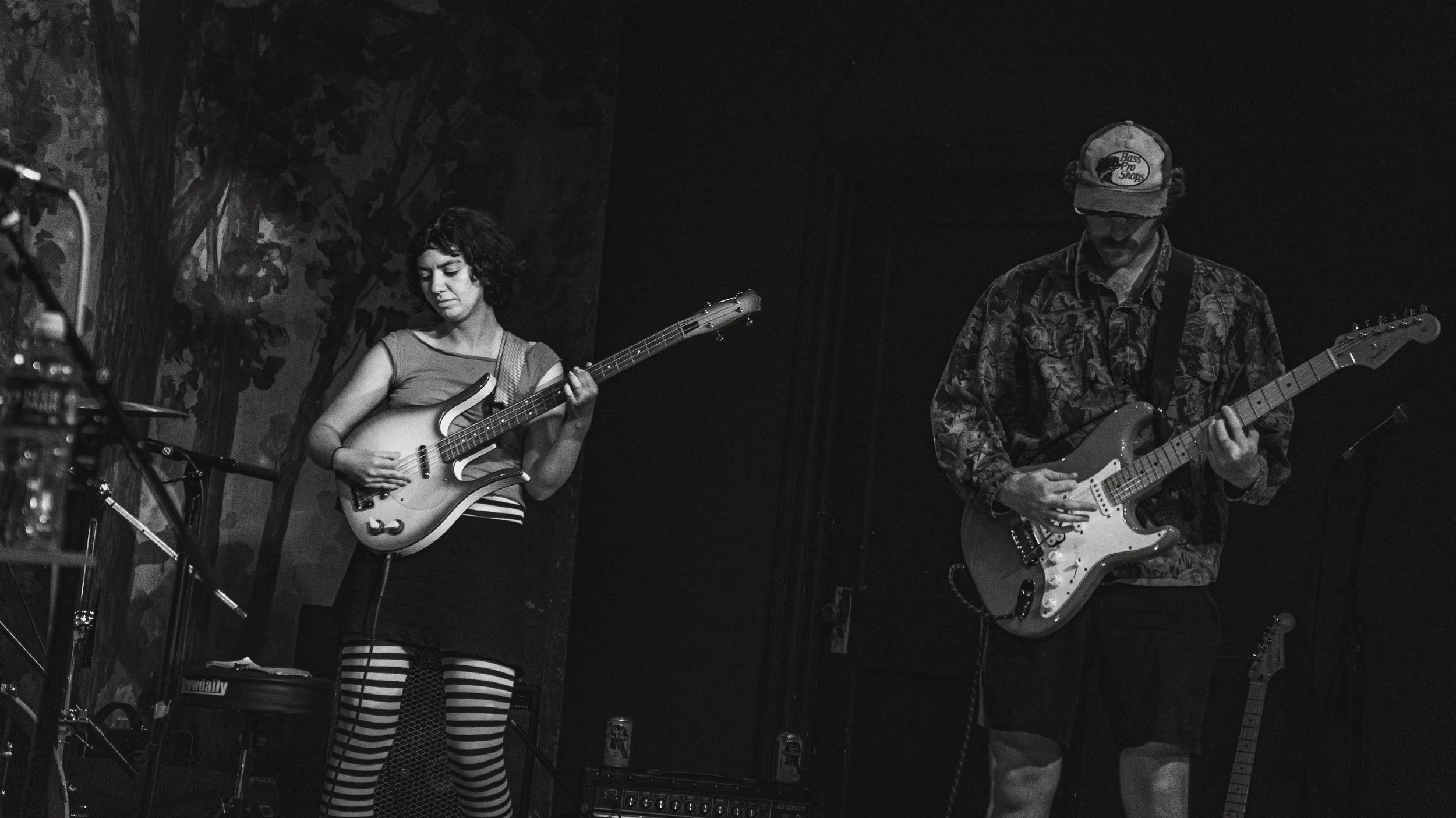 Two musicians playing electric guitars on stage. The woman on the left is wearing striped leggings and a sleeveless top, while the man on the right is wearing a cap and a patterned shirt. The setting appears to be a dimly lit music venue.