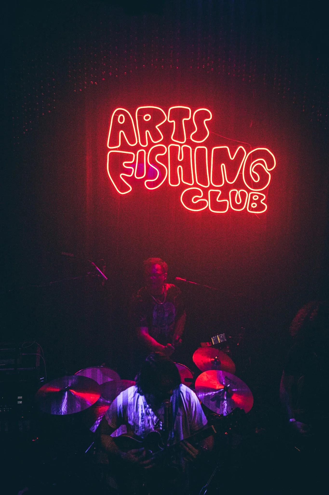 Neon sign reading 'Arts Fushing Club' in red above a drummer playing drums at a musical performance.