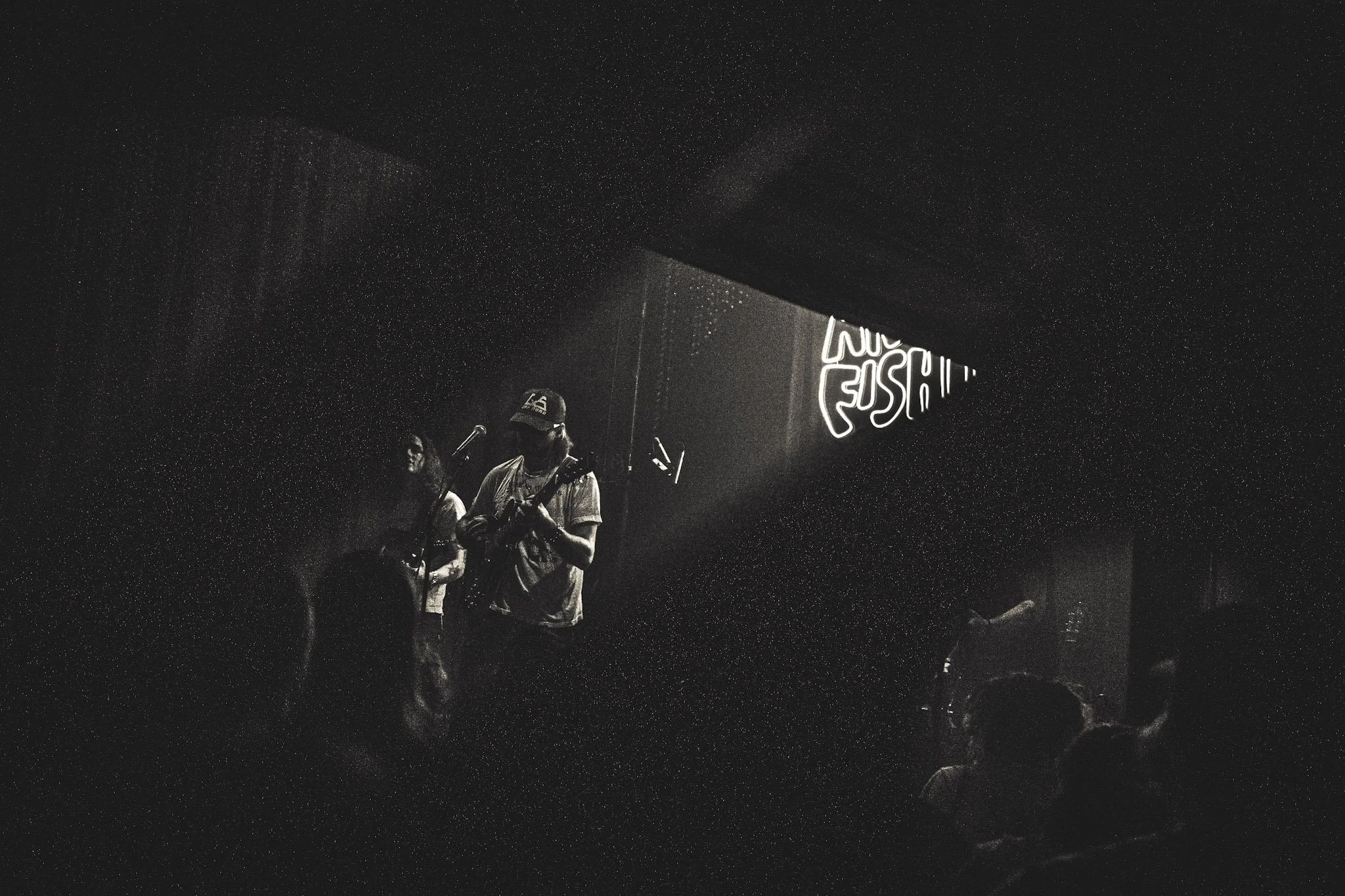 Nighttime scene of a live music performance with band members on stage, illuminated by neon signs, against a starry sky backdrop.