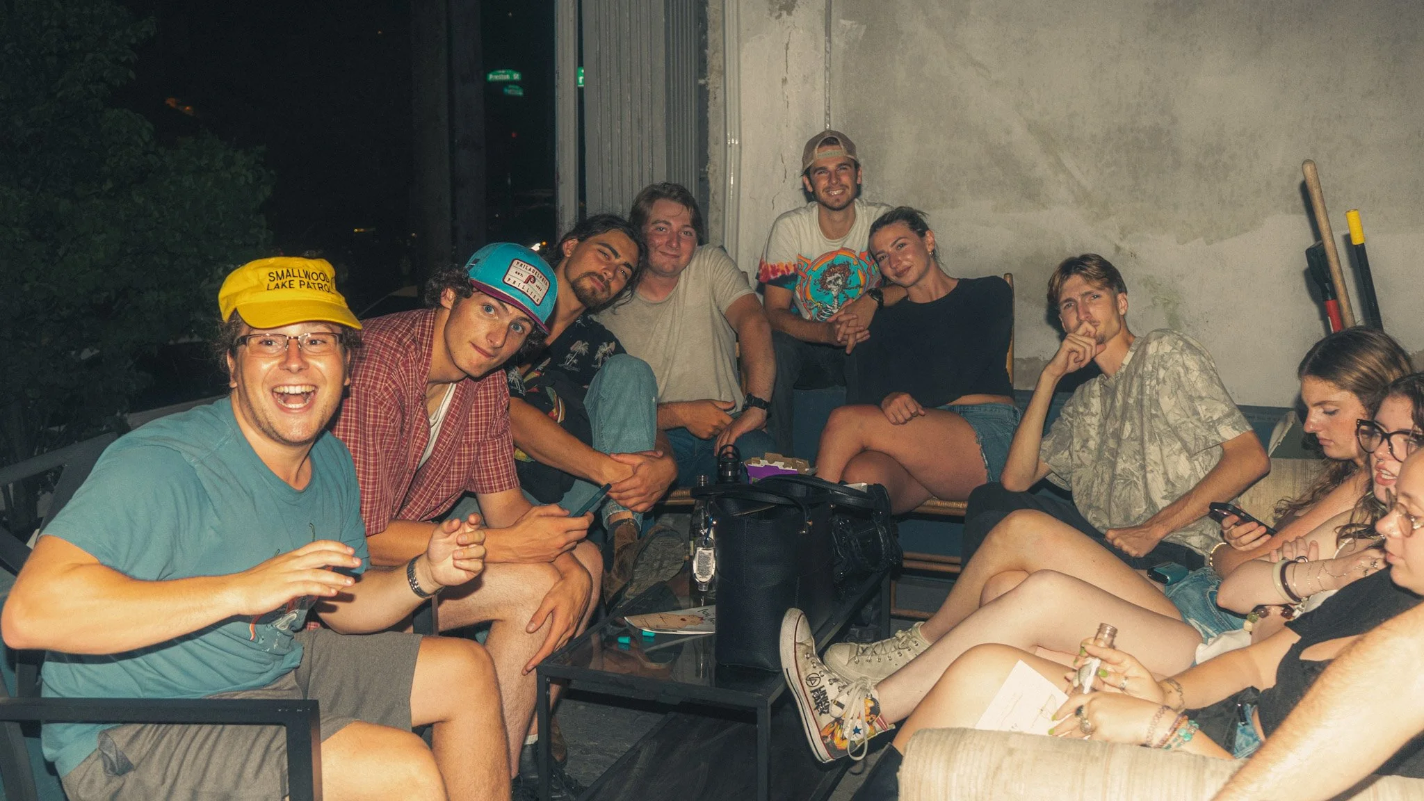 A group of young adults sitting together indoors, smiling, engaging with each other. Some are wearing casual clothing and hats, and there are bottles and personal items on the table in front of them.