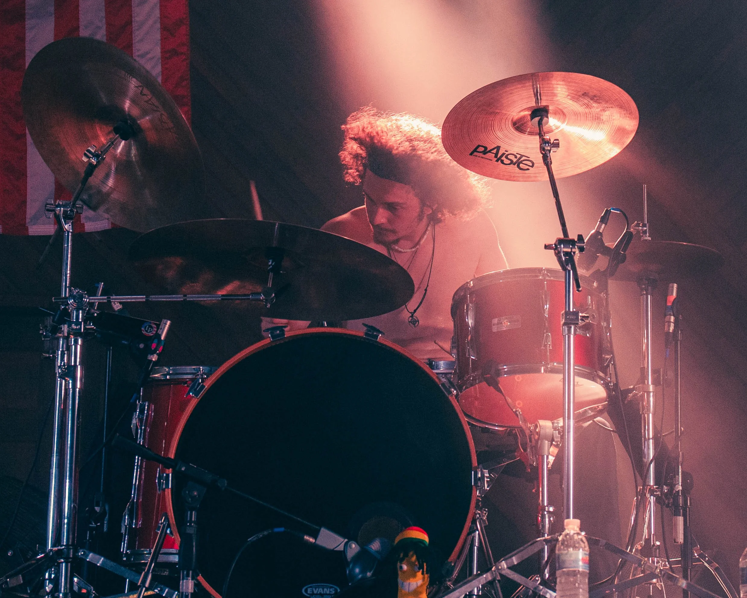 Person playing drums on stage with a large cymbal, stage lighting creating reddish and pinkish hues, water bottle and microphone nearby.