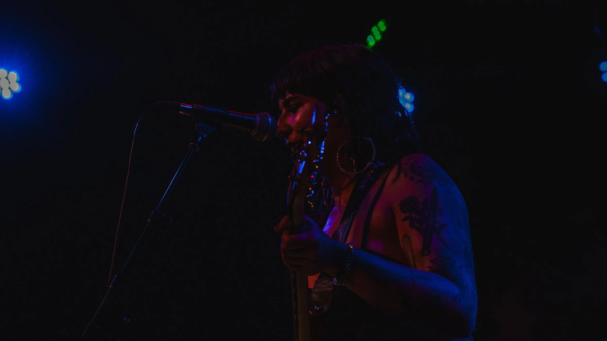A woman singing into a microphone on stage, illuminated by dim, colorful stage lights, with visible tattoos on her arm and hoop earrings.