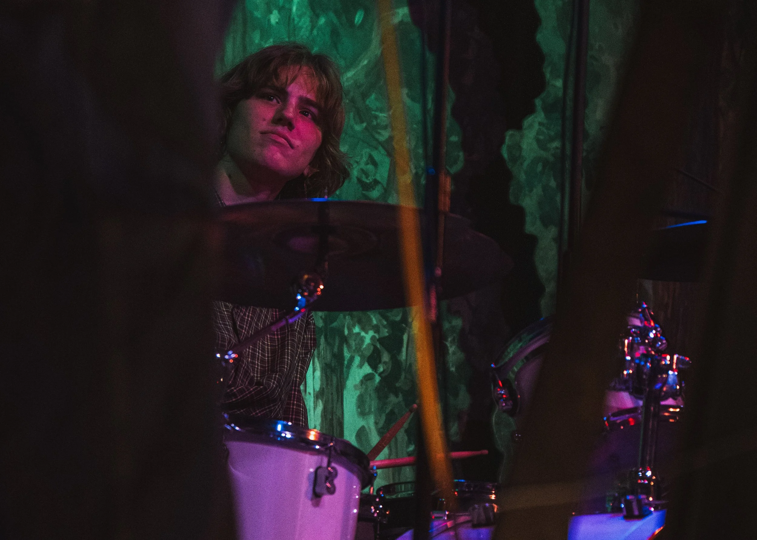 A young person with light brown hair is playing drums behind a drum kit with chrome hardware, illuminated by colorful stage lights. The background features abstract green and blue patterns.