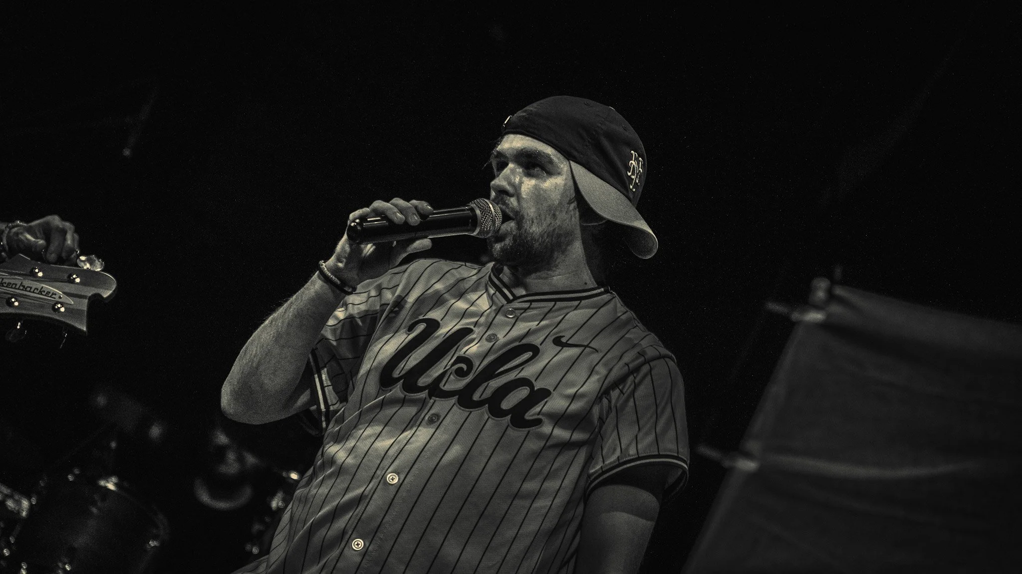 A man with a beard wearing a baseball jersey and a backward cap, holding a microphone while singing or speaking, with a guitar visible on the left side of the image.