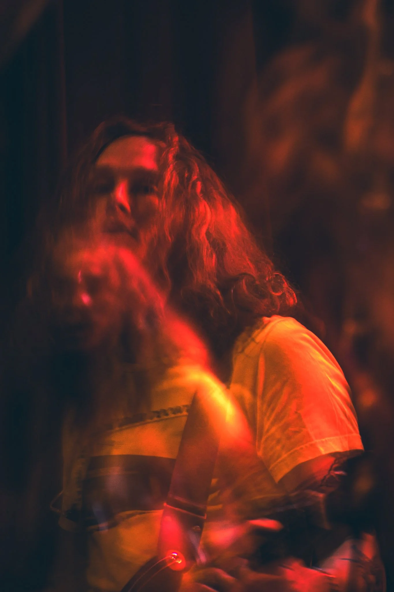 A person with long hair and a beard sitting in a dark room, illuminated by red and orange light. The person is wearing a yellow shirt and glasses, and appears to be looking at the camera.