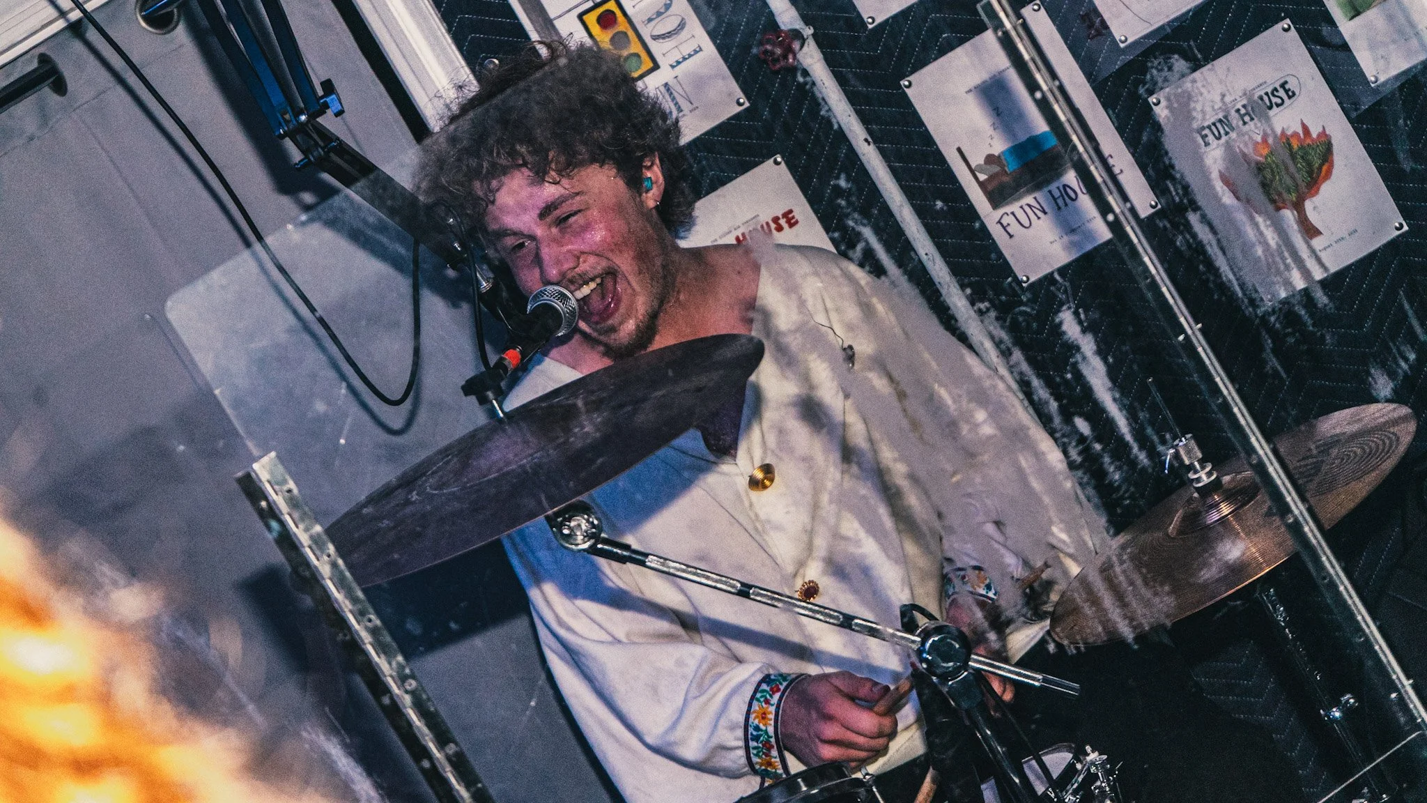 A man with curly hair playing drums, singing into a microphone, with posters in the background that say 'Fun House'.
