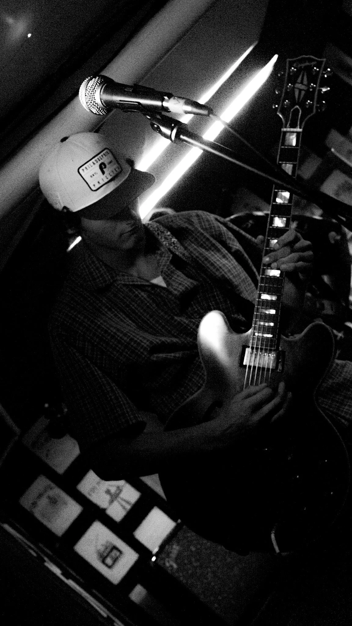 A person wearing a baseball cap and plaid shirt playing an electric guitar in a dimly lit setting, with a microphone in front and a music stand nearby.