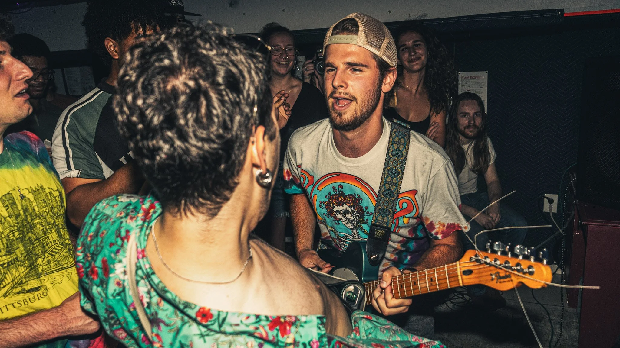 A group of diverse people gathered in a dimly lit room, some smiling and singing, while one person in the foreground plays an acoustic guitar. The atmosphere appears lively and joyful, suggesting a musical or social gathering.