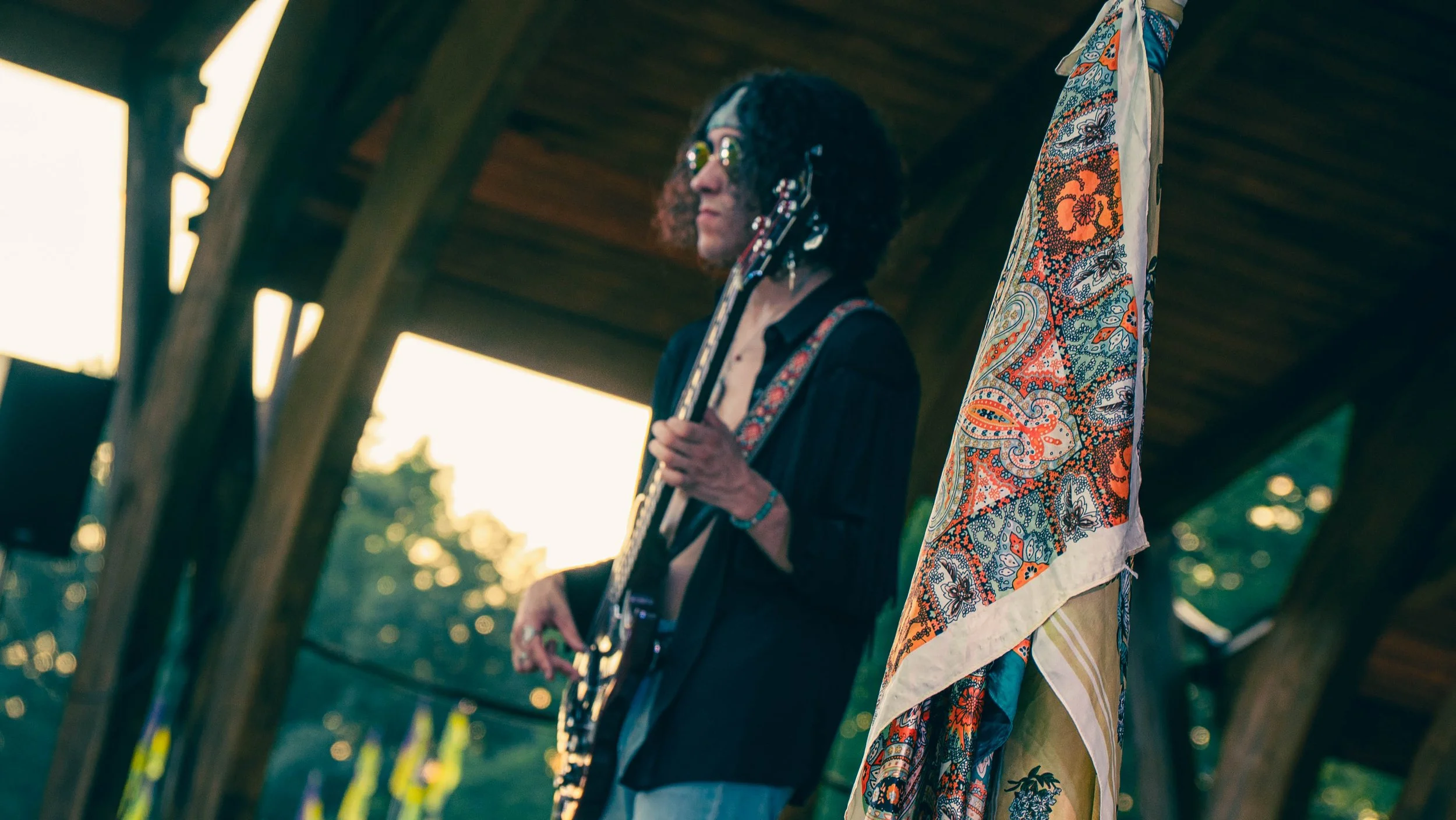 A person with curly hair and glasses playing an electric guitar under a wooden pavilion, with a colorful scarf hanging in the foreground.