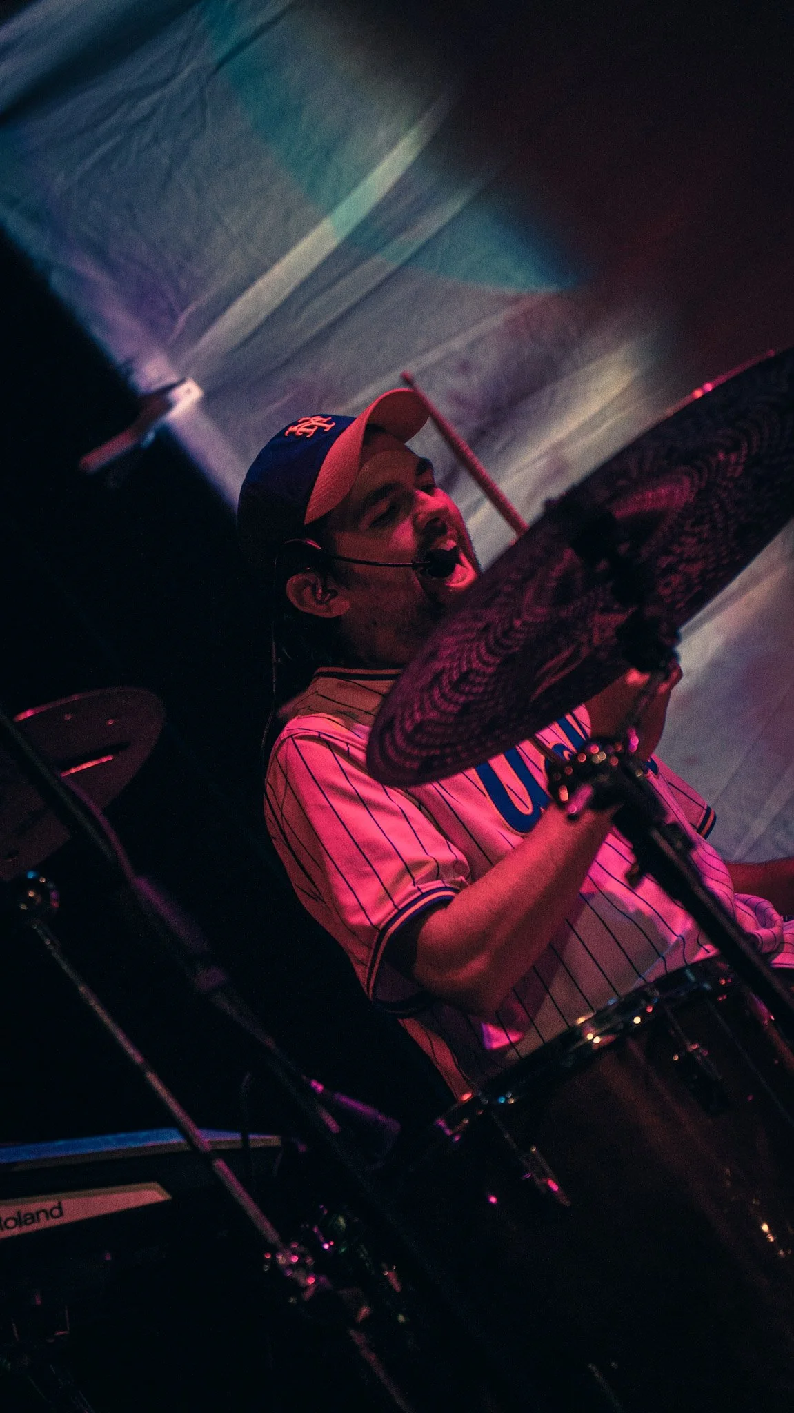 A man singing into a microphone at a DJ setup, wearing a baseball cap, striped shirt, and headphones, with colorful stage lighting in the background.