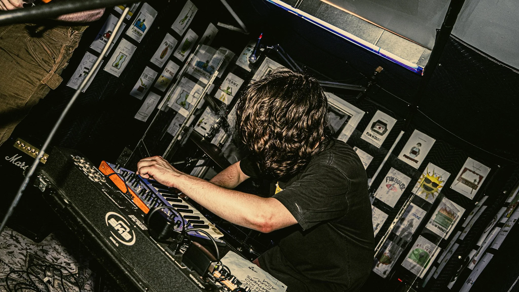 A person with long hair operating synthesizers and electronic music equipment at a music event or studio, with framed artwork and posters on the wall behind.