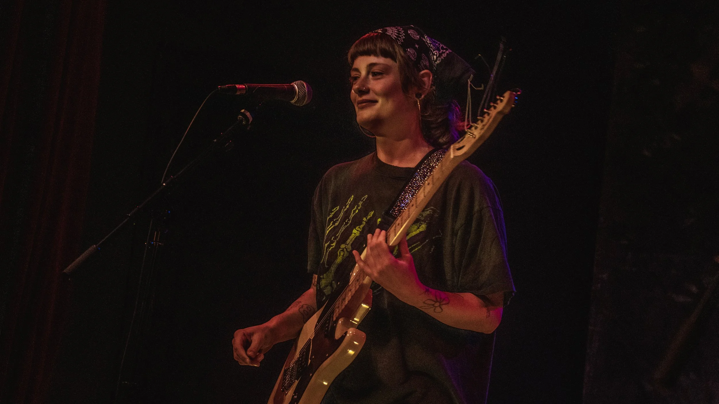 A woman with short hair, tattoos, and a nose ring is playing an electric guitar on stage. She stands in front of a microphone, smiling softly. The scene is dimly lit with stage lights.