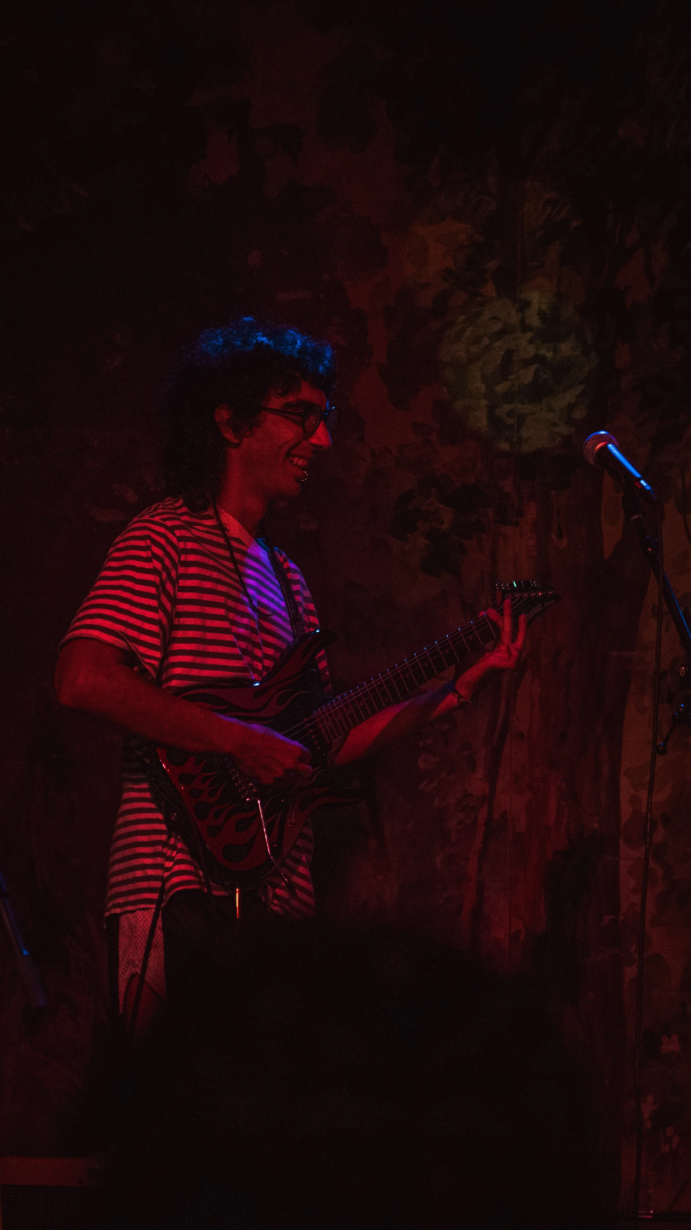 A person with curly hair and glasses playing an electric guitar on stage, smiling, wearing a striped shirt, with a microphone nearby, in a dimly lit venue with a dark patterned backdrop.