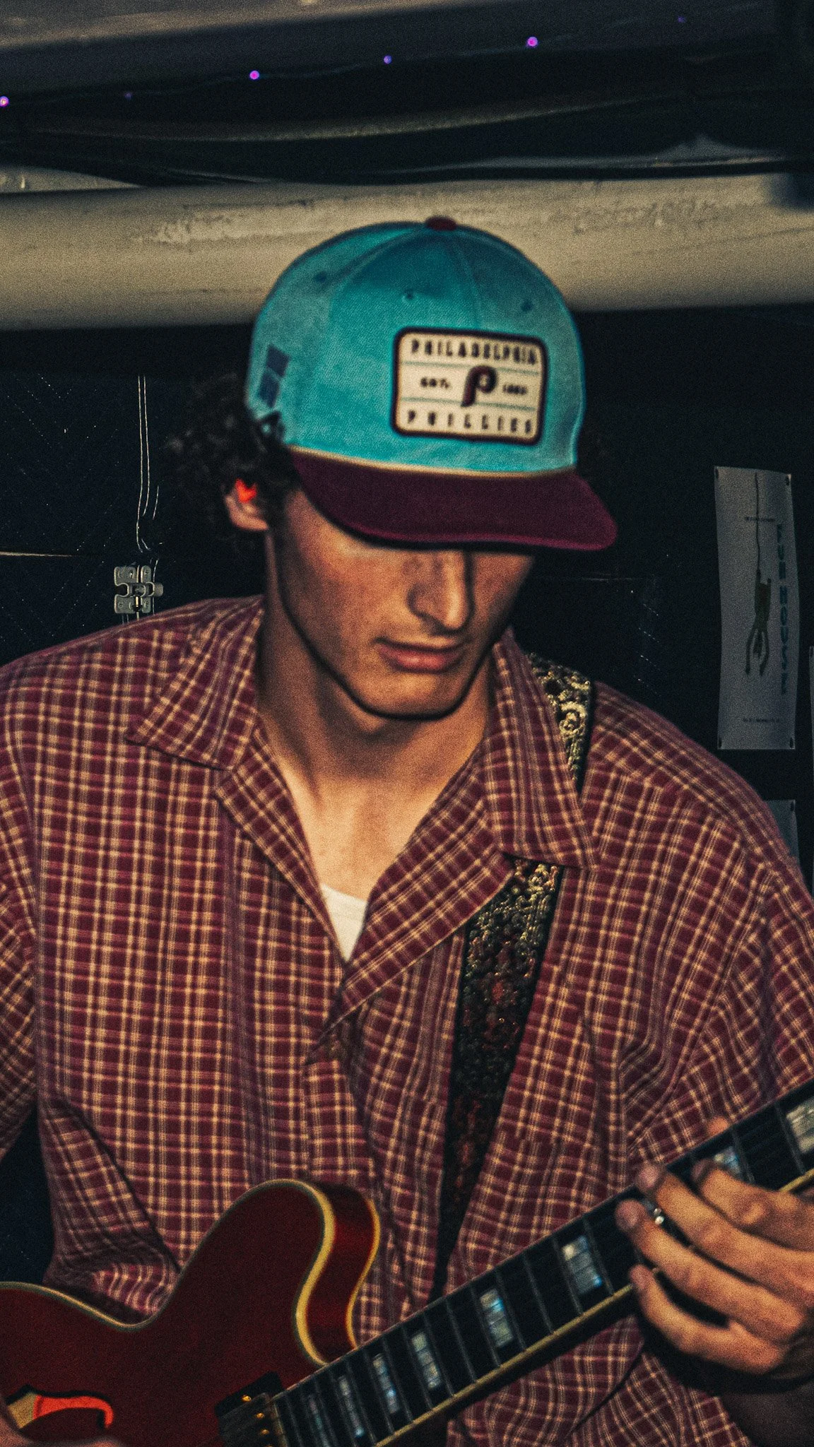 A young man wearing a red and teal baseball cap with a graphic patch, a plaid shirt, and playing an electric guitar.