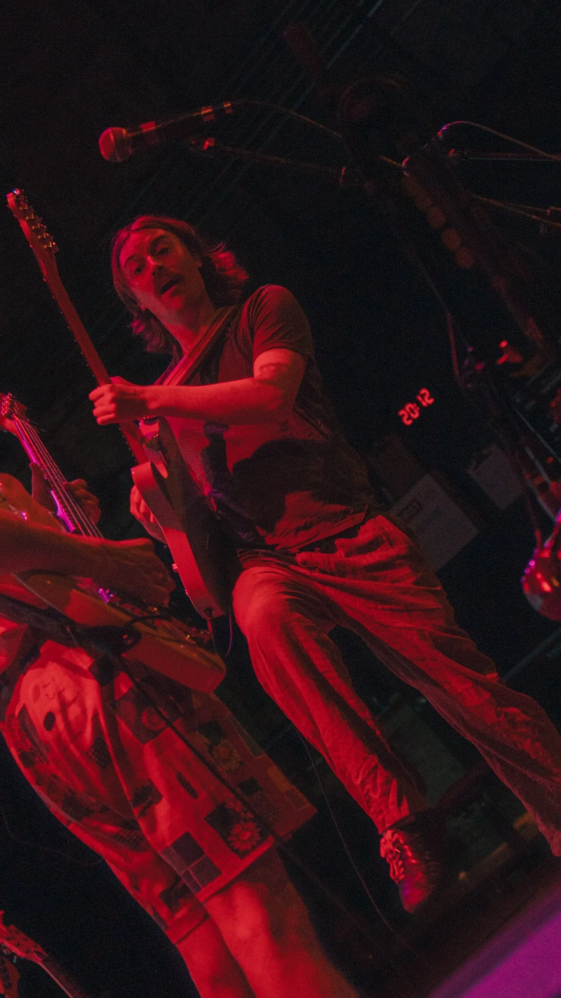 A man playing an electric guitar under red lighting, with a reflection on a shiny surface, and audio equipment nearby.