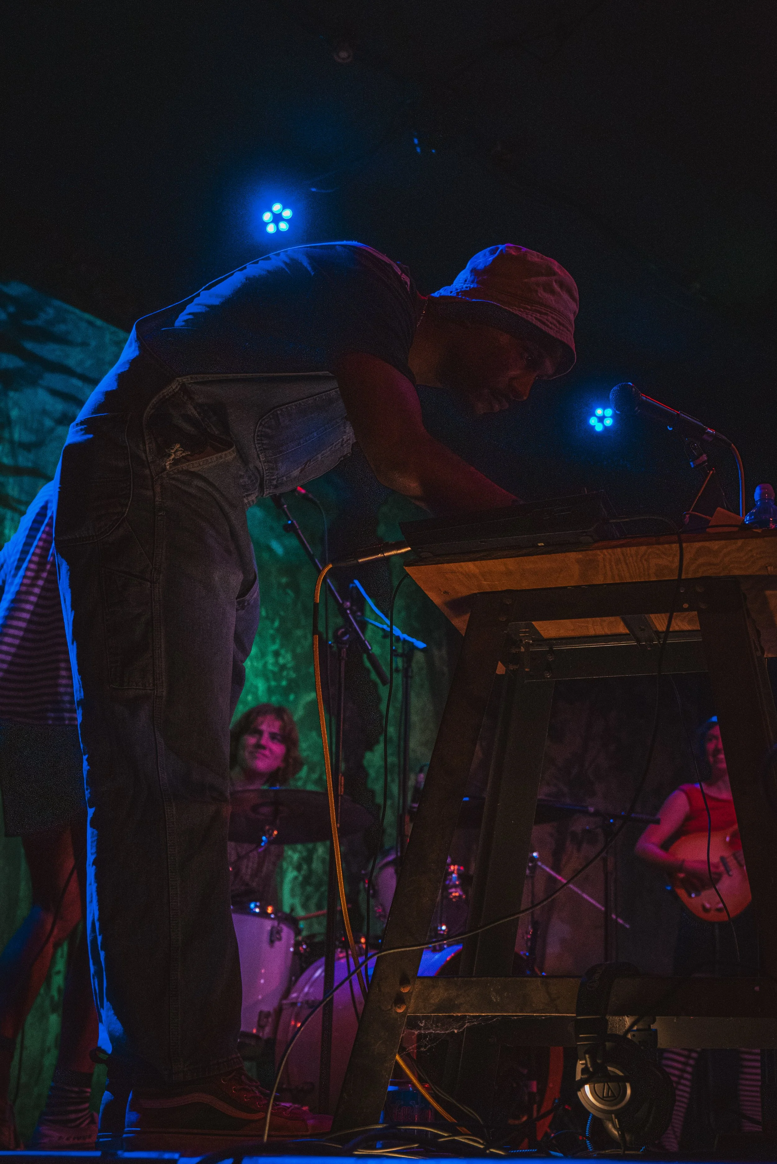 A man wearing a hoodie and jeans performing with electronic music equipment on stage. There are people in the background, including a woman with a guitar. The scene is lit with colorful stage lights.
