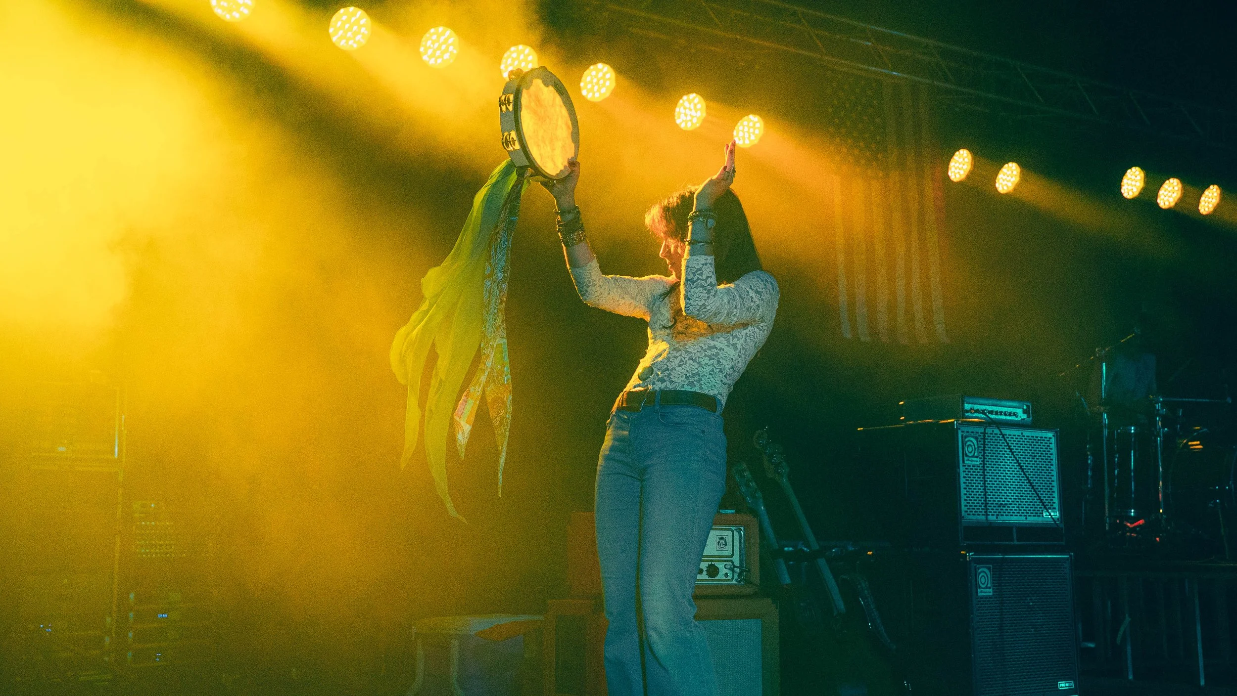 A person on stage holding a tambourine and performing in front of bright yellow stage lights, with musical equipment in the background.