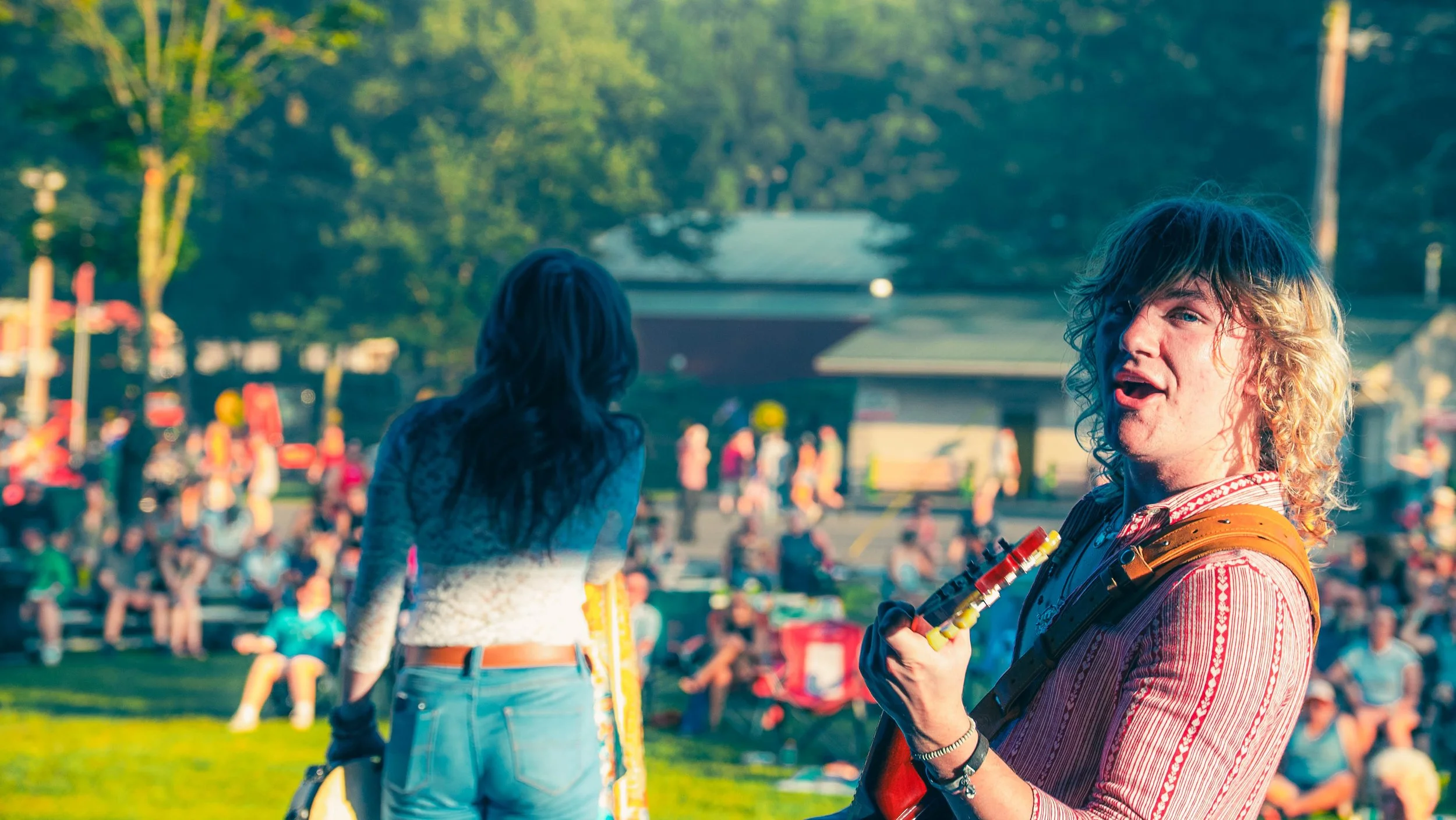 A young man with long, curly blond hair and wearing a striped shirt is holding a colorful toy or gadget at an outdoor event with many people and trees in the background.