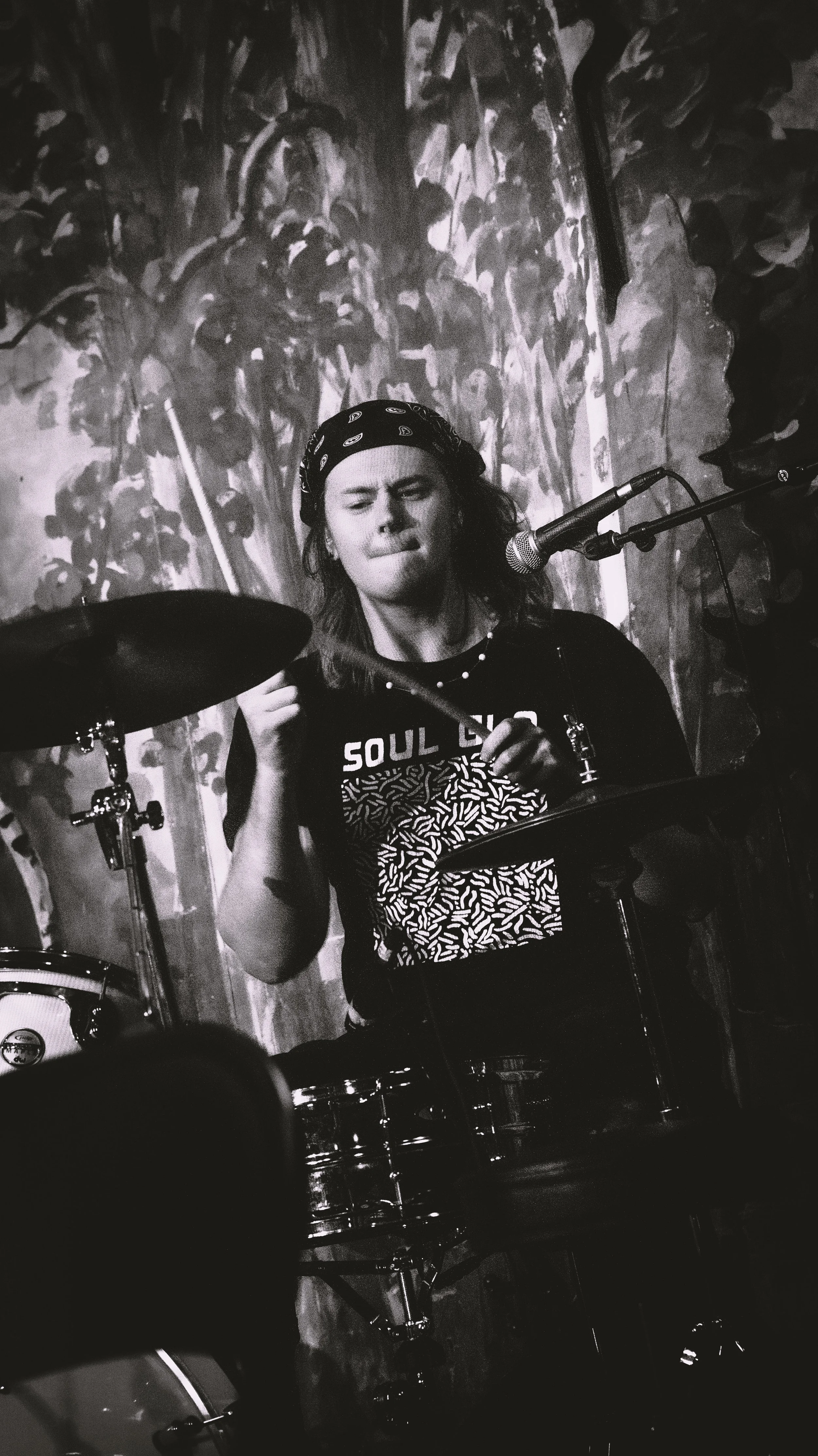 A person with long hair, wearing a black bandana and a black shirt with a graphic design, is playing drums on stage, singing into a microphone. The stage has a textured painting or mural in the background.