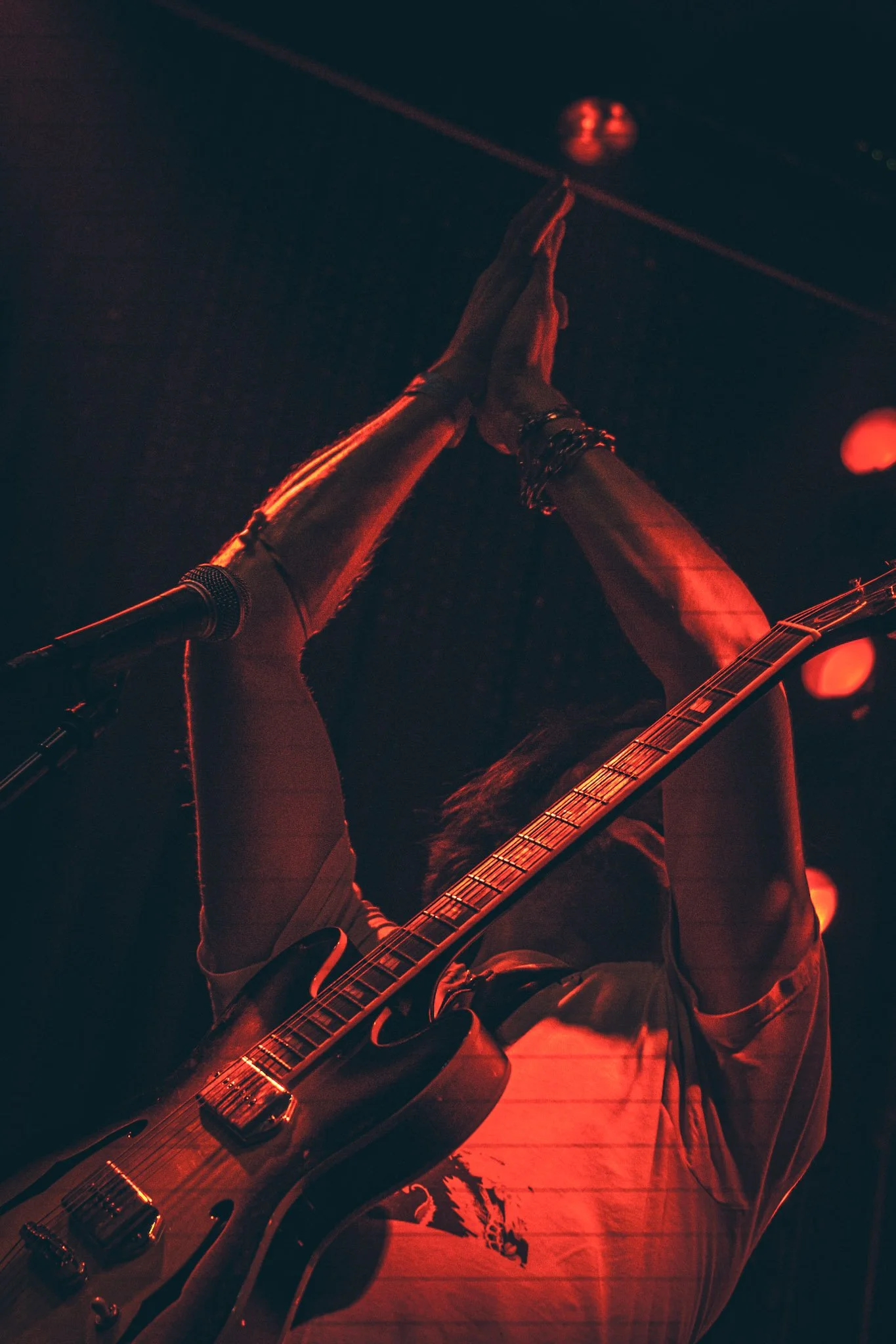A person playing an electric guitar on stage with red lighting, their arms raised and hands pressed together in prayer gestures, wearing bracelets and a striped shirt.