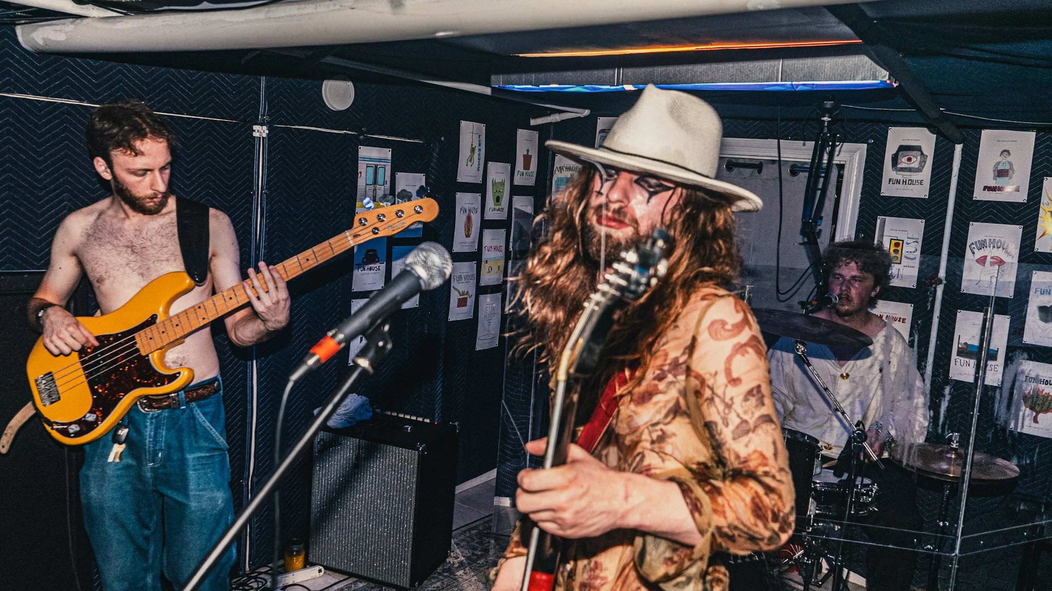 A band performs live in a small venue with three members visible: a shirtless guitarist with long curly hair, a woman with long hair and a wide-brimmed hat playing guitar, and a drummer with curly hair behind a drum set. The venue has walls decorated