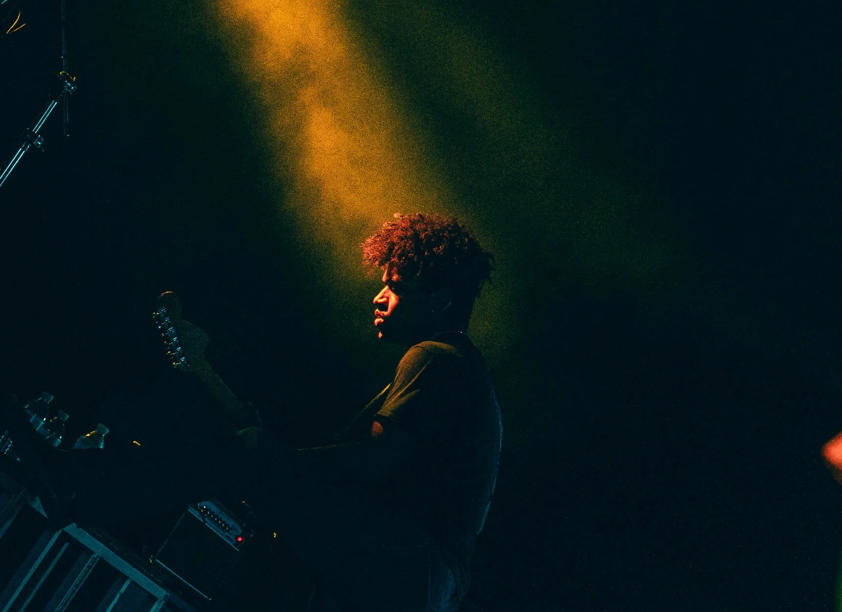 A person with curly hair holding a guitar on stage with dark background and yellowish lighting.