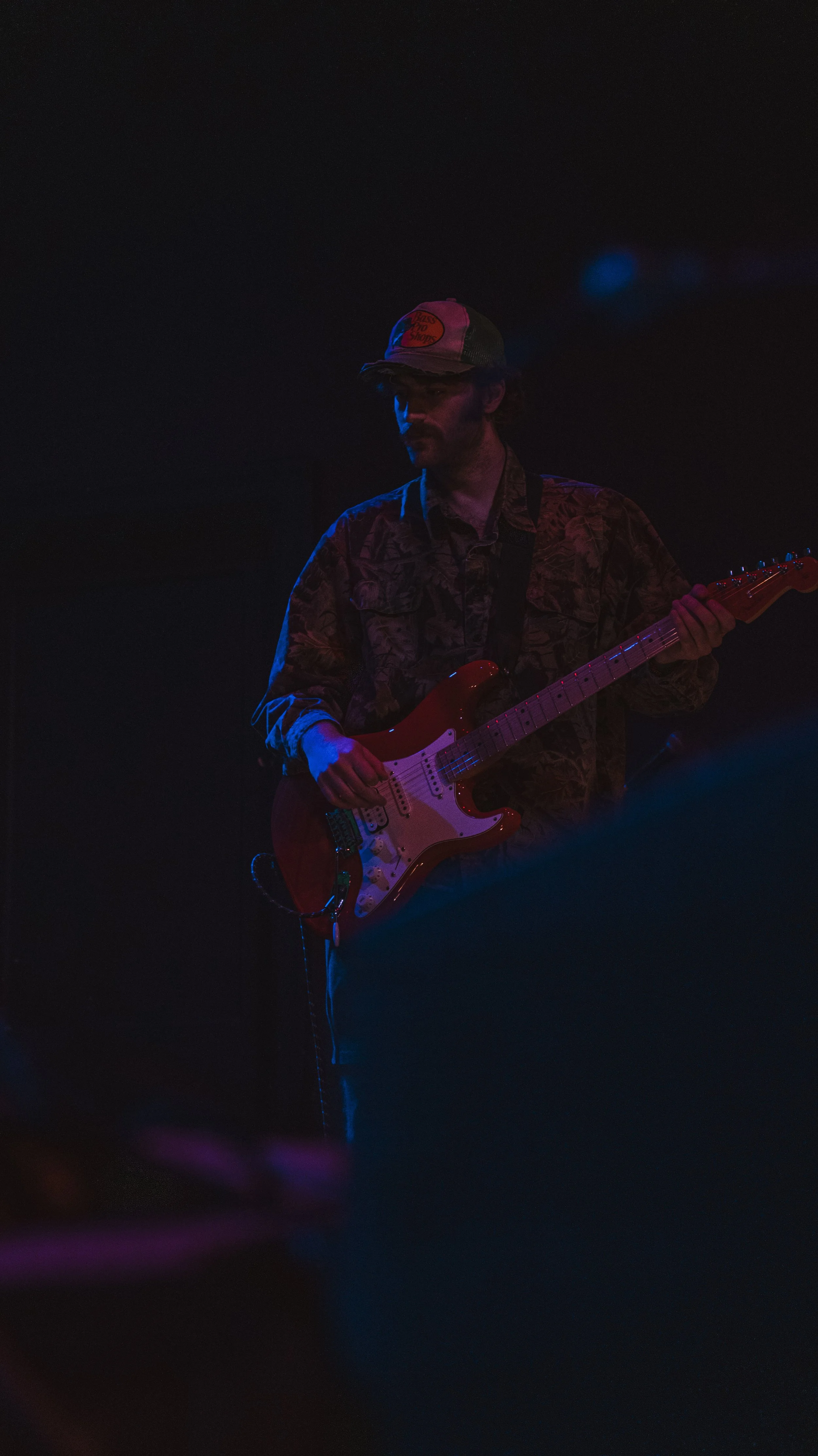 A man wearing a baseball cap and patterned shirt playing an electric guitar on stage under low lighting.