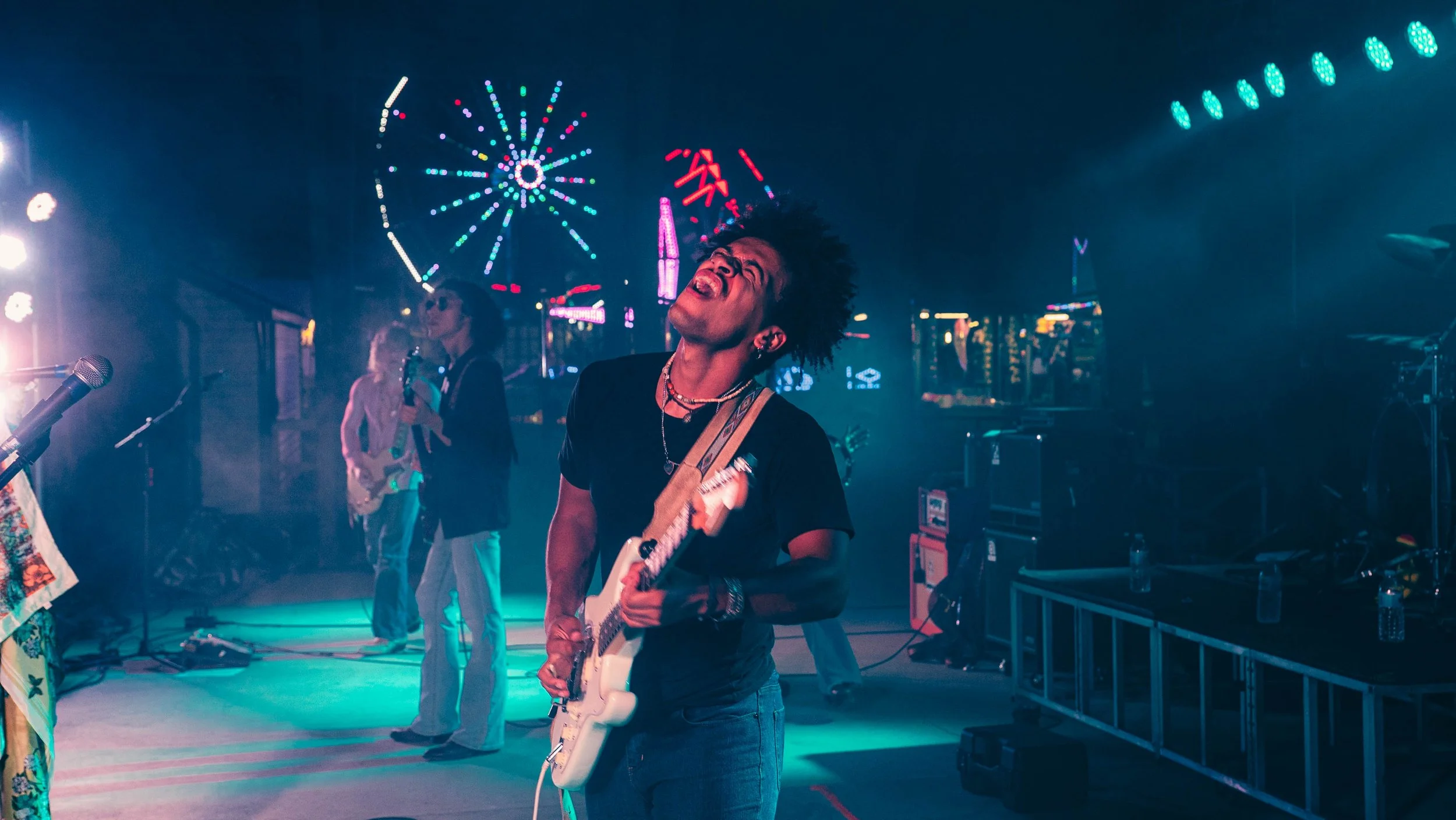 A person with curly hair, wearing a black shirt and a necklace, plays an electric guitar with their eyes closed and mouth open, at a nighttime outdoor event with colorful Ferris wheel and amusement park rides in the background.