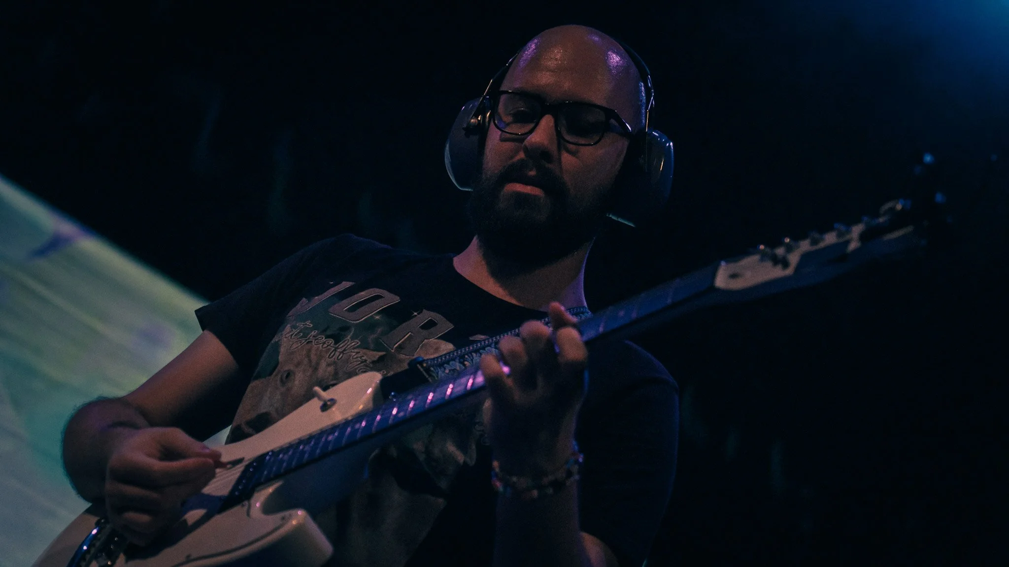 A man with glasses and a beard playing an electric guitar in a dimly lit setting, with purple and blue lighting.