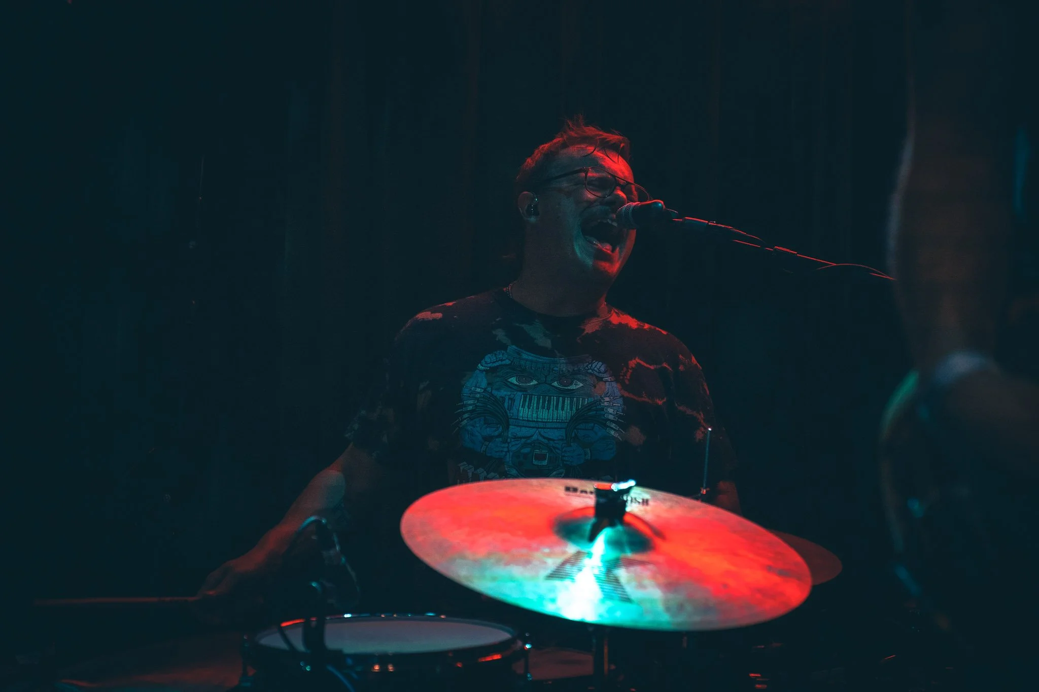 A musician wearing glasses and a graphic t-shirt is performing with a drum set under red and blue stage lighting.
