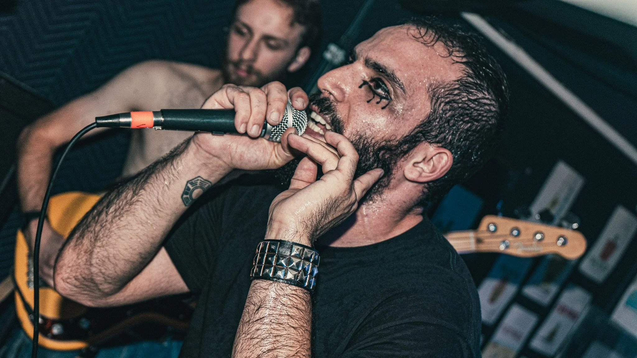 A man singing into a microphone with a guitar in the background, another person resting nearby, both in a music performance setting.