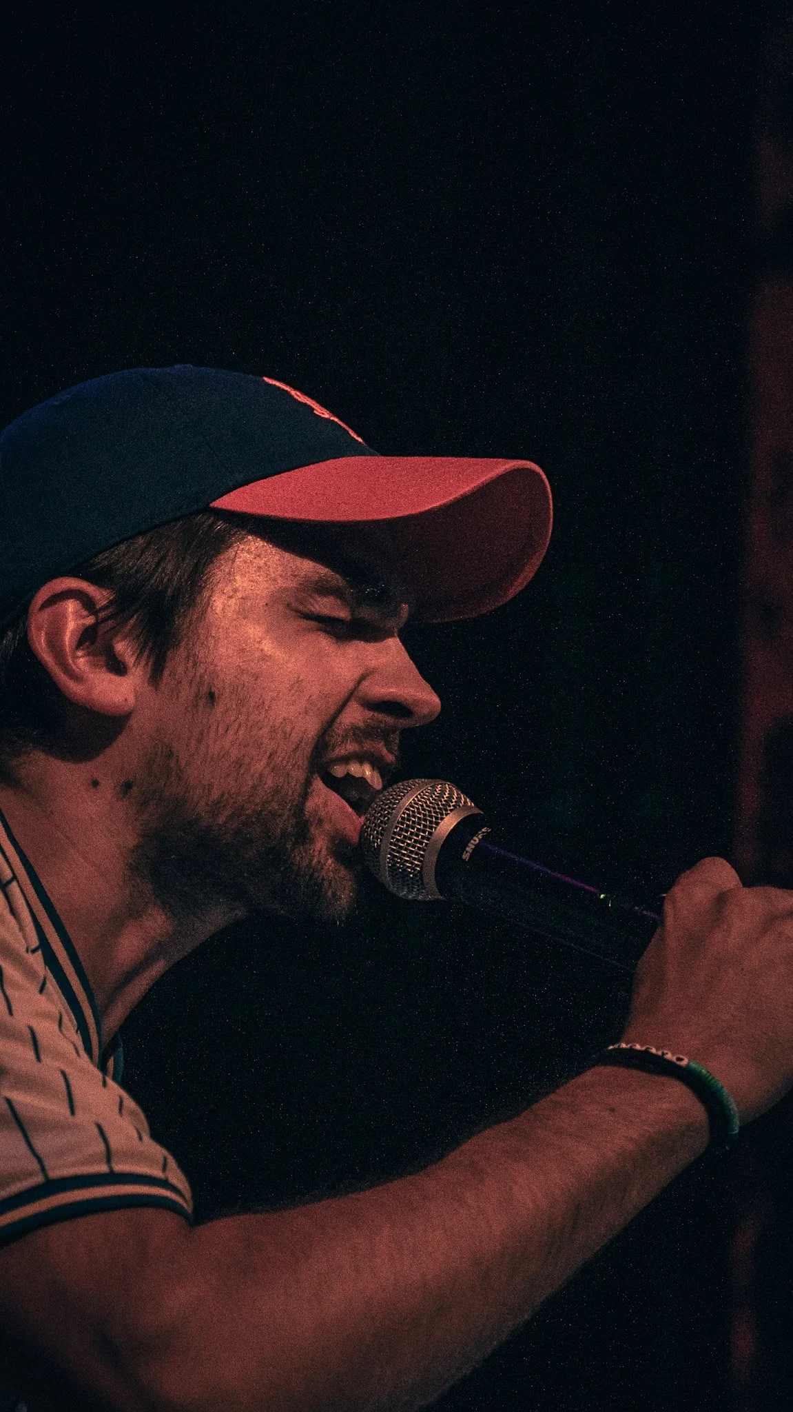 A man singing into a microphone, wearing a blue and red baseball cap, with a nighttime background filled with stars.