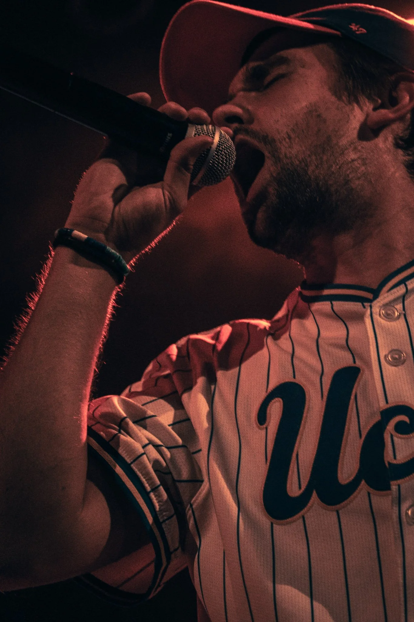 A man singing into a microphone with his eyes closed, wearing a baseball jersey with black and white vertical stripes and a number 36, and a cap. The lighting is dim and red-toned.