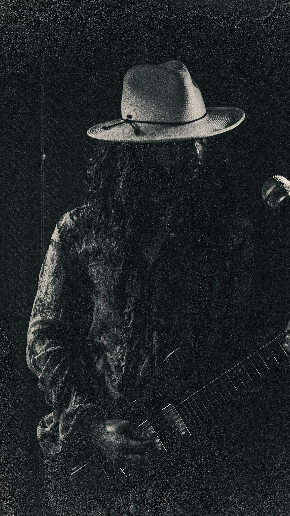 A person with long hair wearing a cowboy hat, patterned shirt, and necklace, holding a guitar in a dark, textured room.