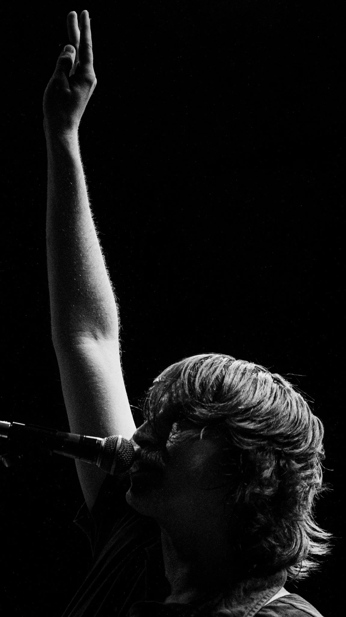 Black and white photo of a woman with styled hair raising her left arm, with a microphone positioned near her face.