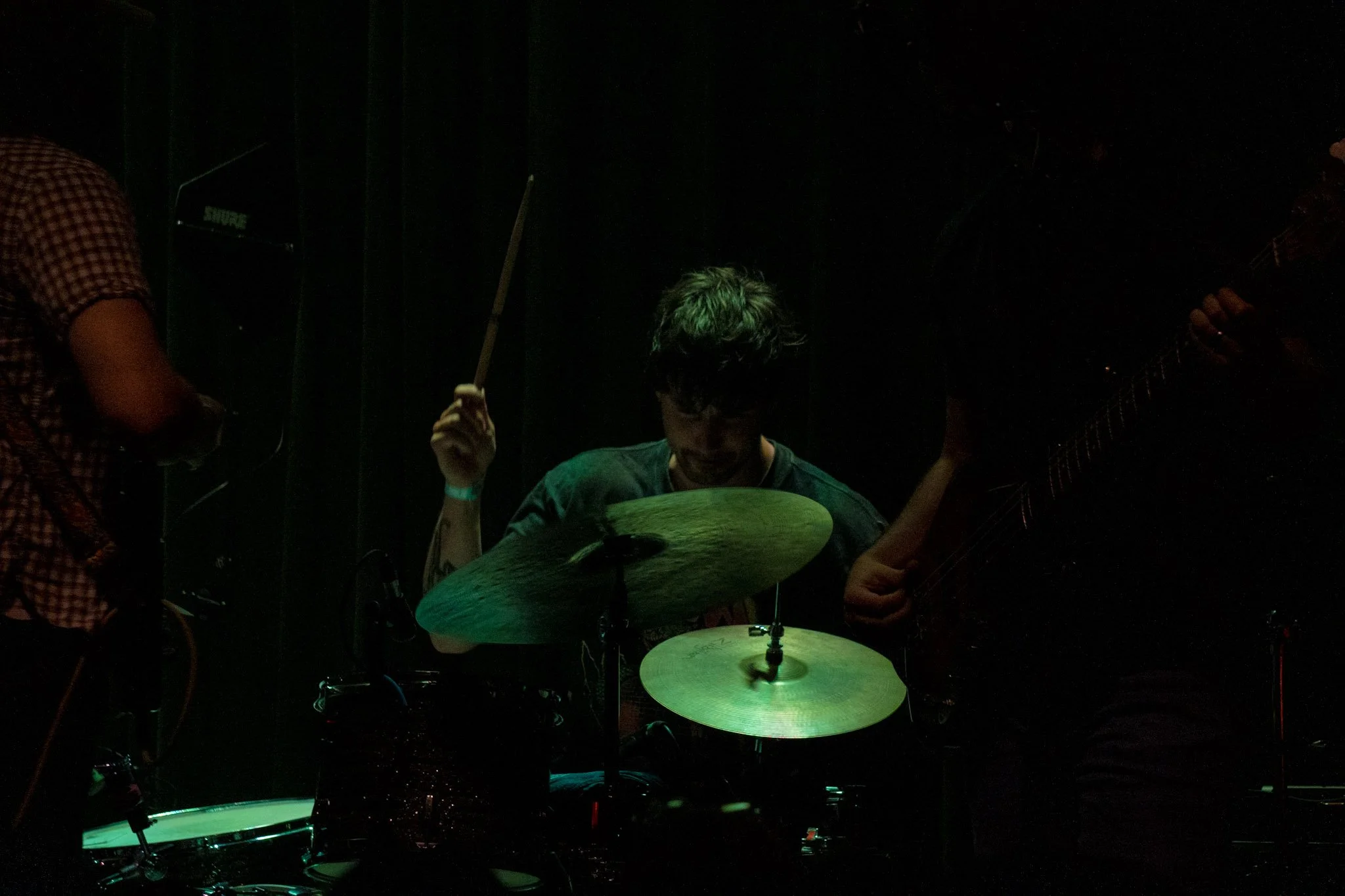 A dimly lit scene of musicians, including a drummer with green-highlighted hair and another person holding a guitar, performing on stage.