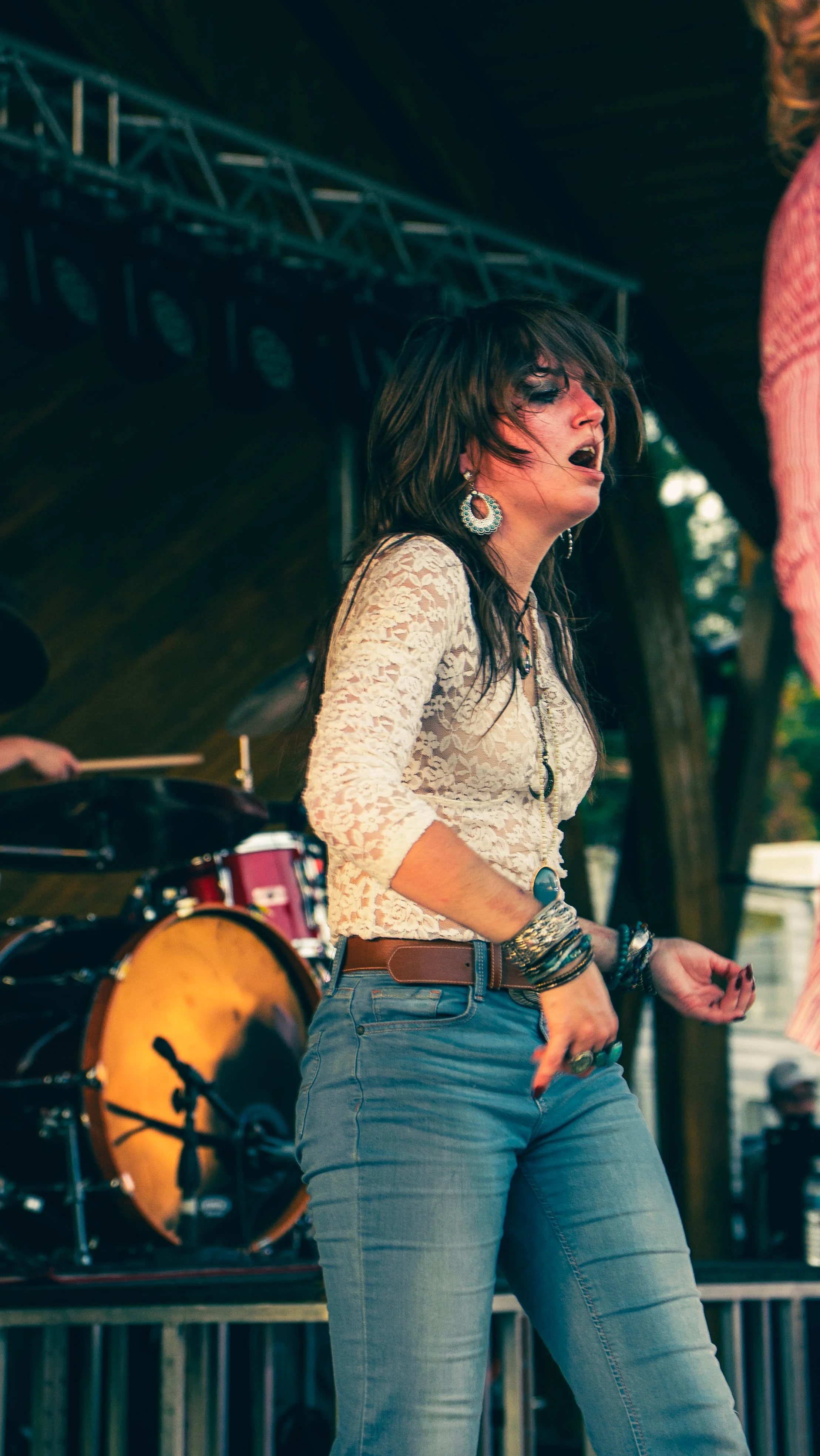 A woman with long dark hair, wearing a lacy white top, blue jeans, and large earrings, appears to be singing or performing at an outdoor music event. There is a drum set behind her and a stage structure overhead.