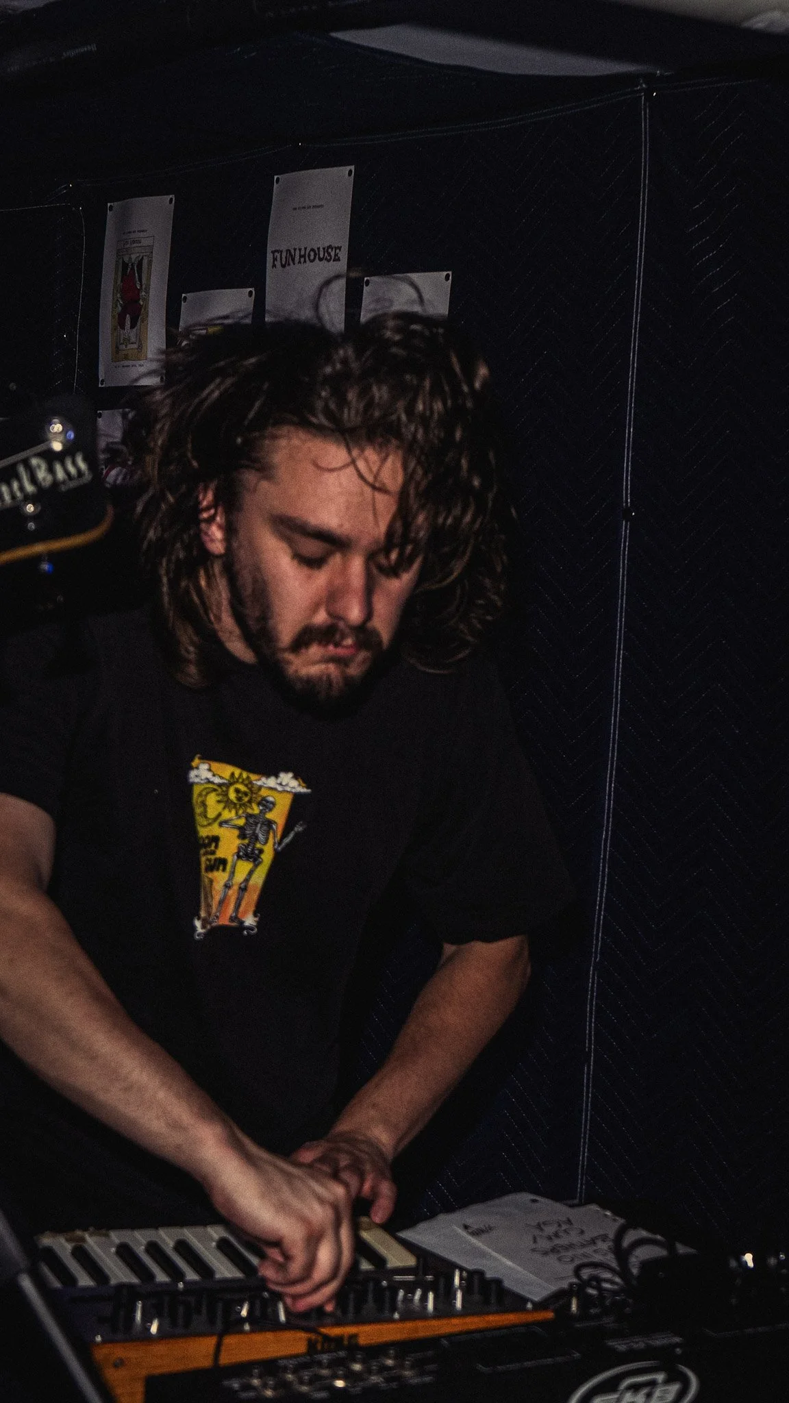 A man with long curly hair and a beard playing electronic music on a keyboard, wearing a black T-shirt with a skeleton graphic, in a dimly lit indoor setting.
