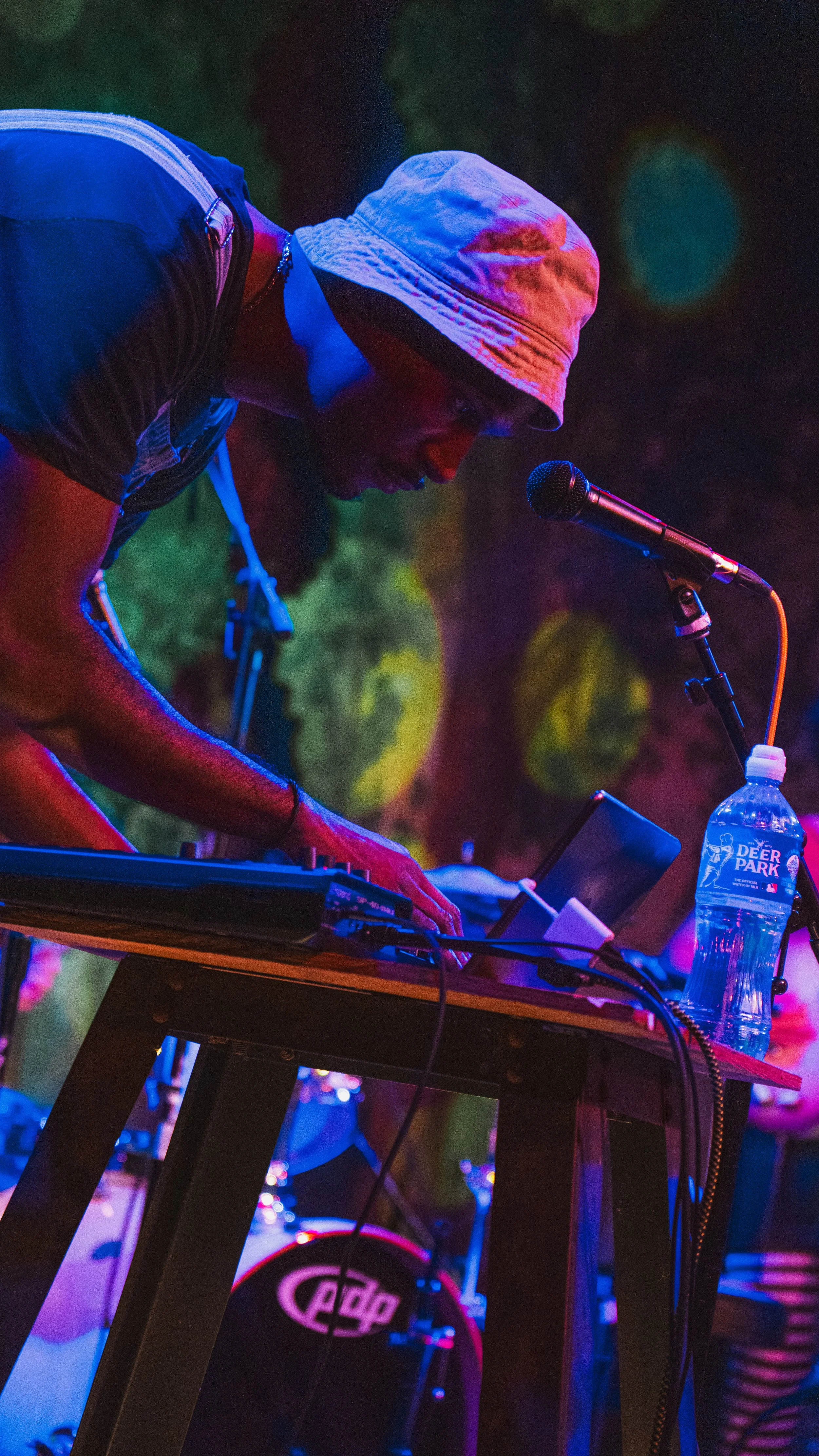 A person performing electronic music on stage, wearing a light-colored bucket hat and dark clothing, with a microphone and music equipment in front of them, colorful stage lighting in the background.