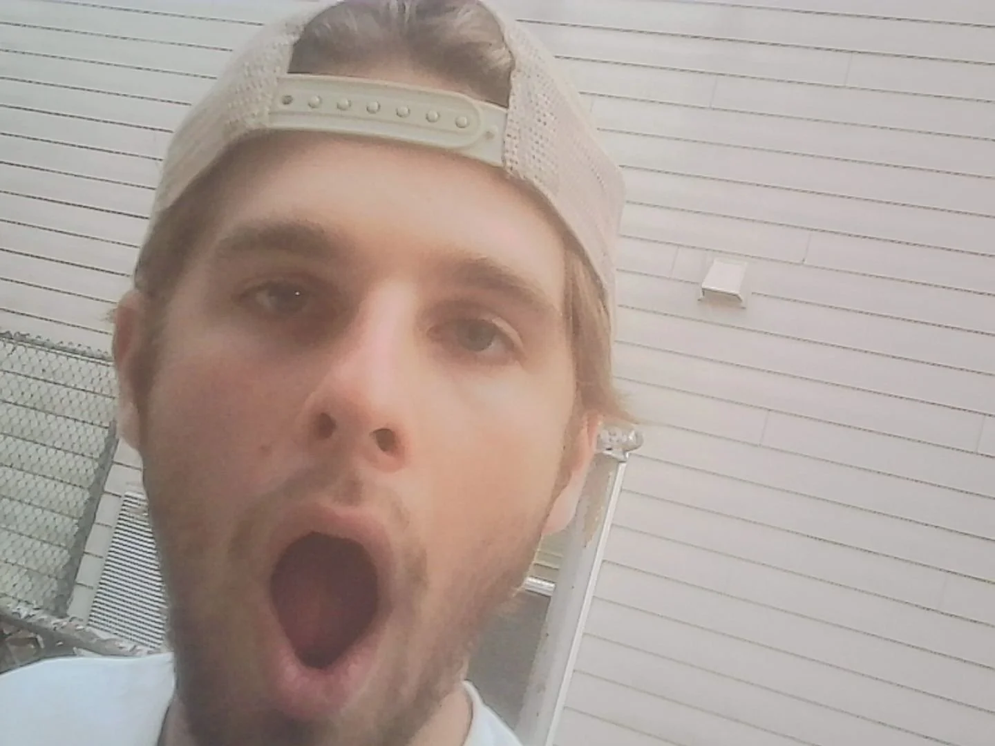 Young man wearing a baseball cap backward with mouth open in surprise or amazement, outdoor setting with white siding wall and metal fence in background.