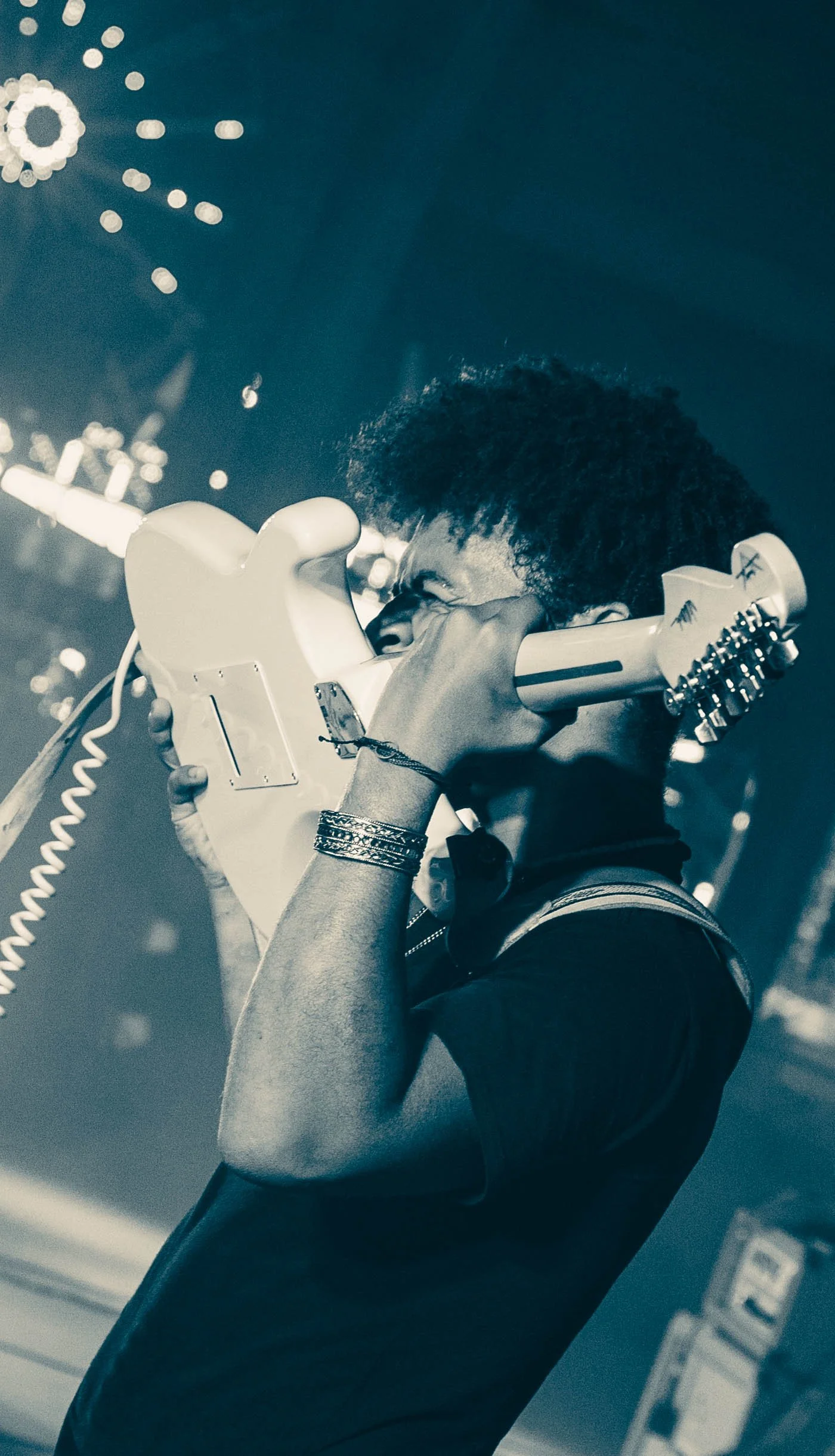 A person with curly hair playing an electric guitar, captured in black and white, wearing bracelets on wrist, closed eyes, intense expression, stage lighting and blurred background.