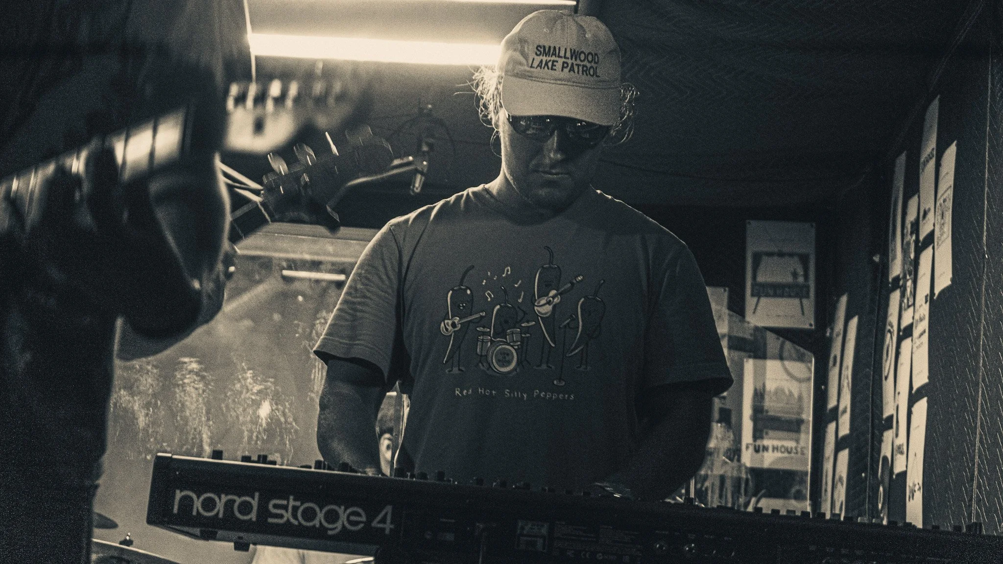 A person wearing a baseball cap and sunglasses standing behind a keyboard, with a sign reading 'Nord Stage 4' and posters or signs on the wall behind him.