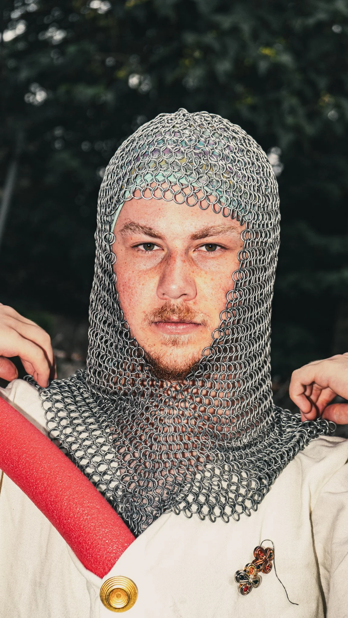 A person wearing chainmail armor on their head and neck, holding a red foam tube, with a background of dark green foliage.