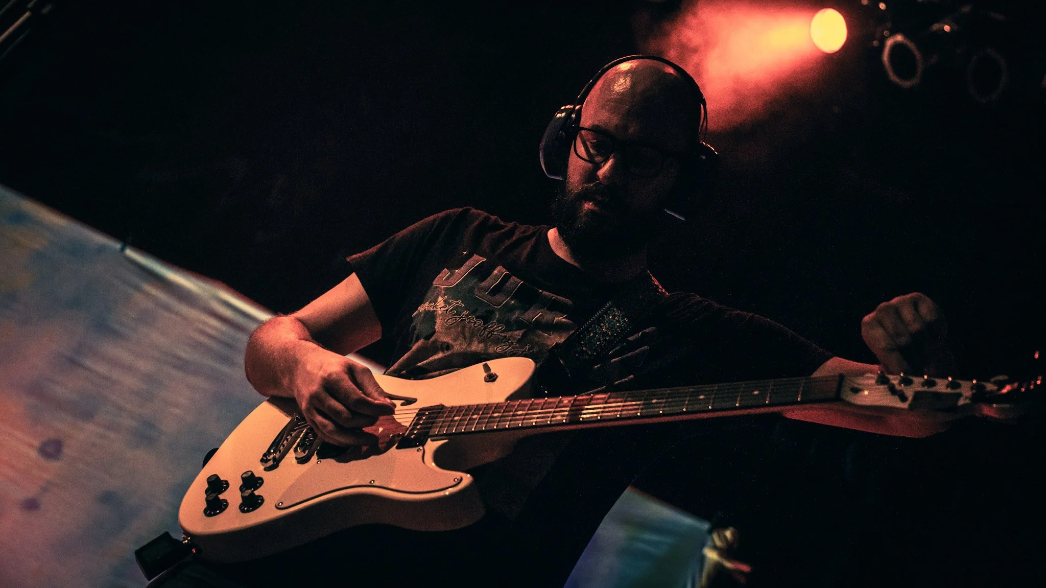 A man with glasses and a beard playing an electric guitar on stage, with red stage lights illuminating the background.