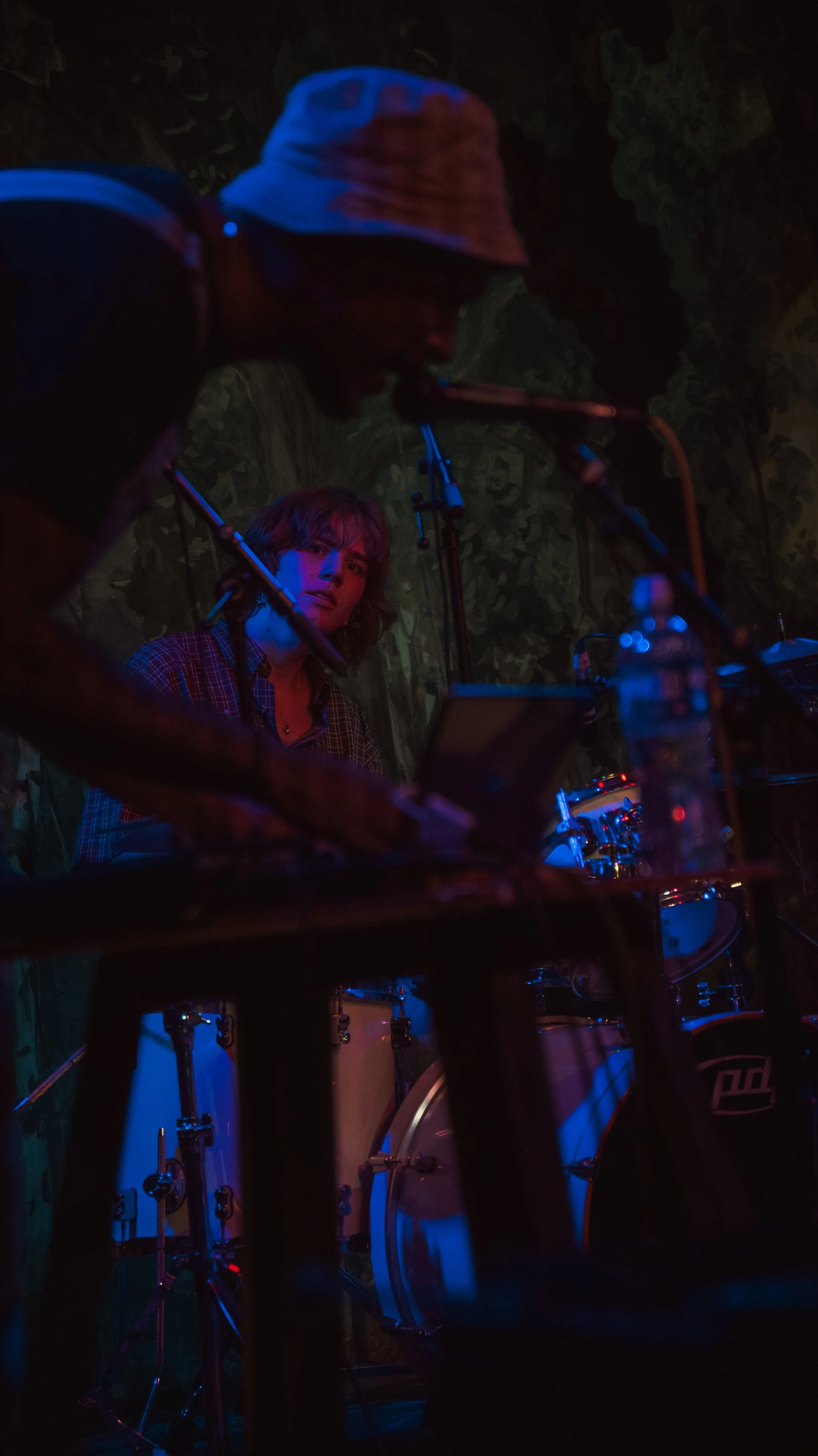 A person wearing a bucket hat and glasses is playing drums on stage, with another person singing or speaking into a microphone. The scene is dimly lit with blue and purple lighting, and there is a leafy background.