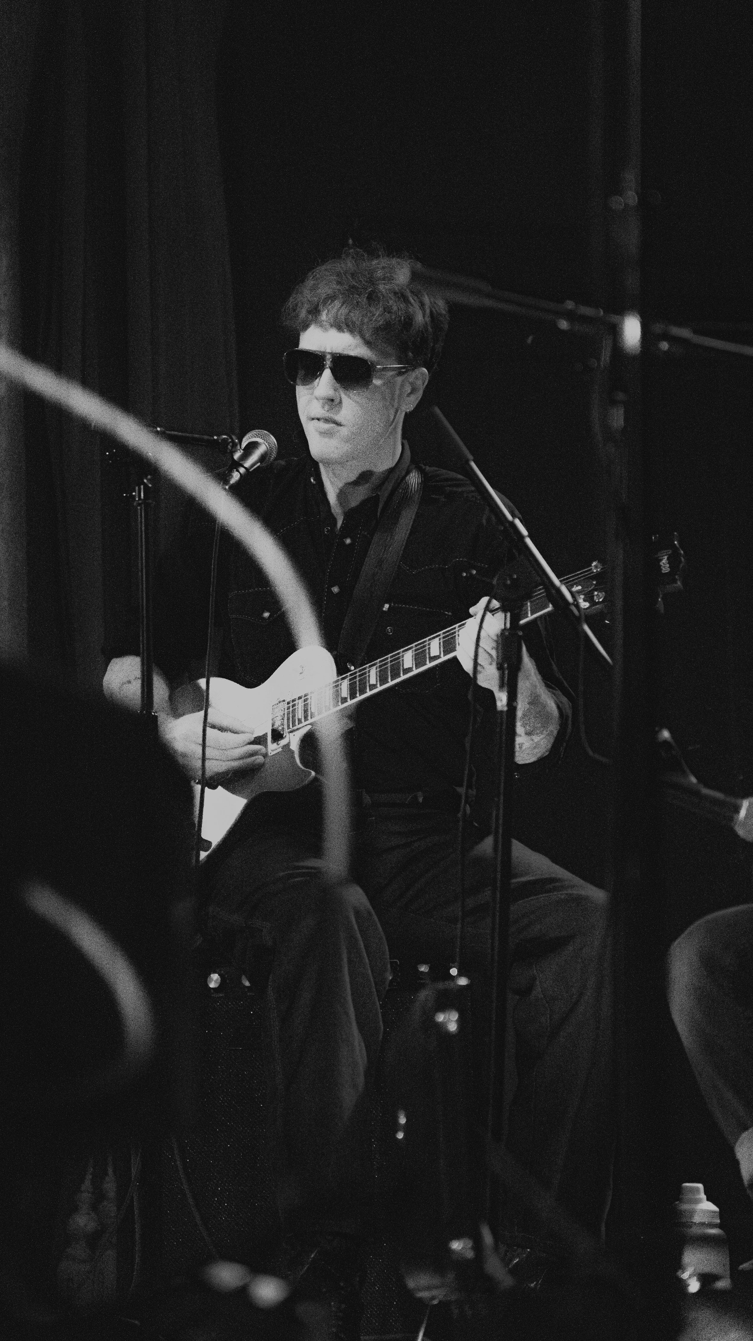 A person wearing sunglasses and a dark shirt playing an acoustic guitar in front of a microphone during a performance or recording session in a dark room.