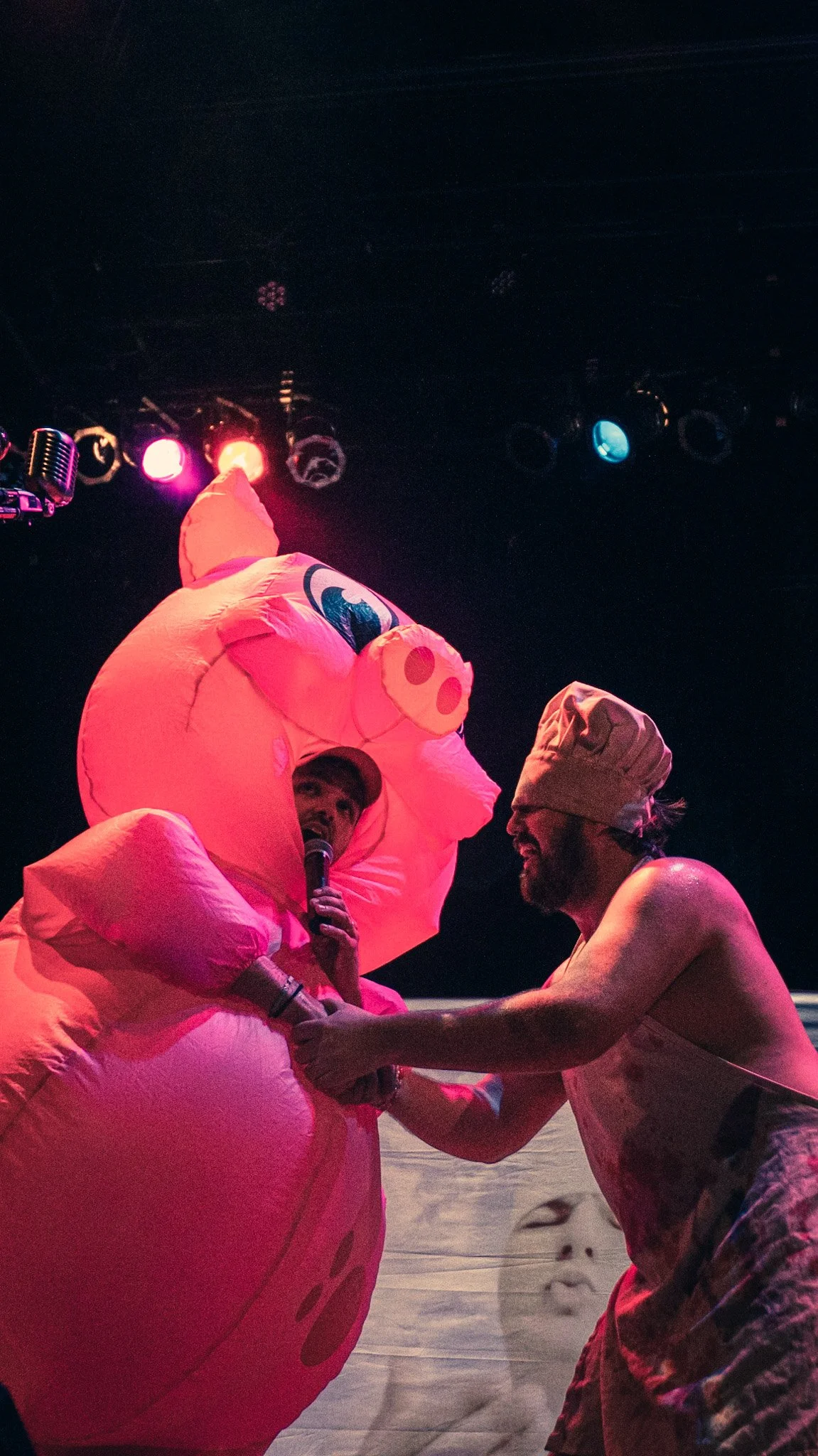Two men are on stage, one dressed in a pink pig costume and the other shirtless with a bandana, holding hands. The man in the pig costume is speaking into a microphone, and there are colorful stage lights overhead.