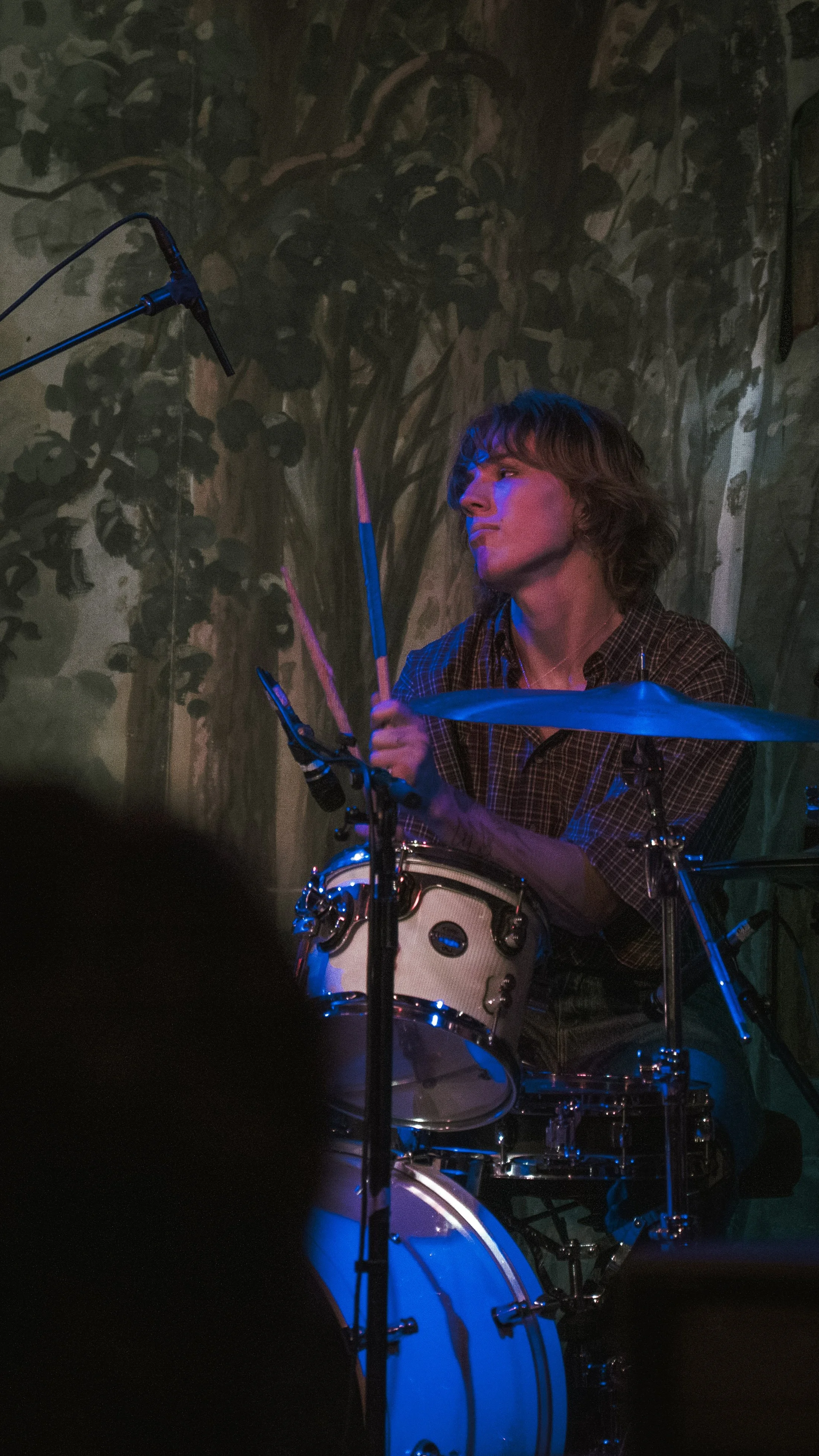 A woman with short, wavy hair plays the drums on stage, with a colorful mural of trees painted on the wall behind her. She appears focused and engaged in her performance, with a microphone positioned nearby.