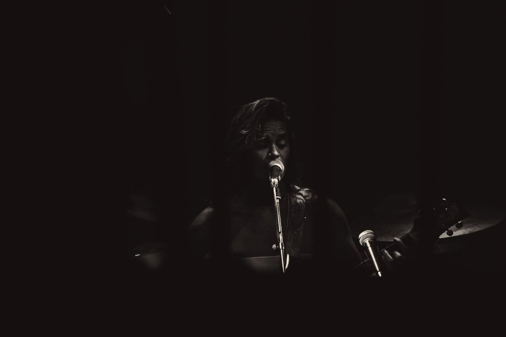 A woman singing into a microphone with a dark background, accompanied by a person playing guitar